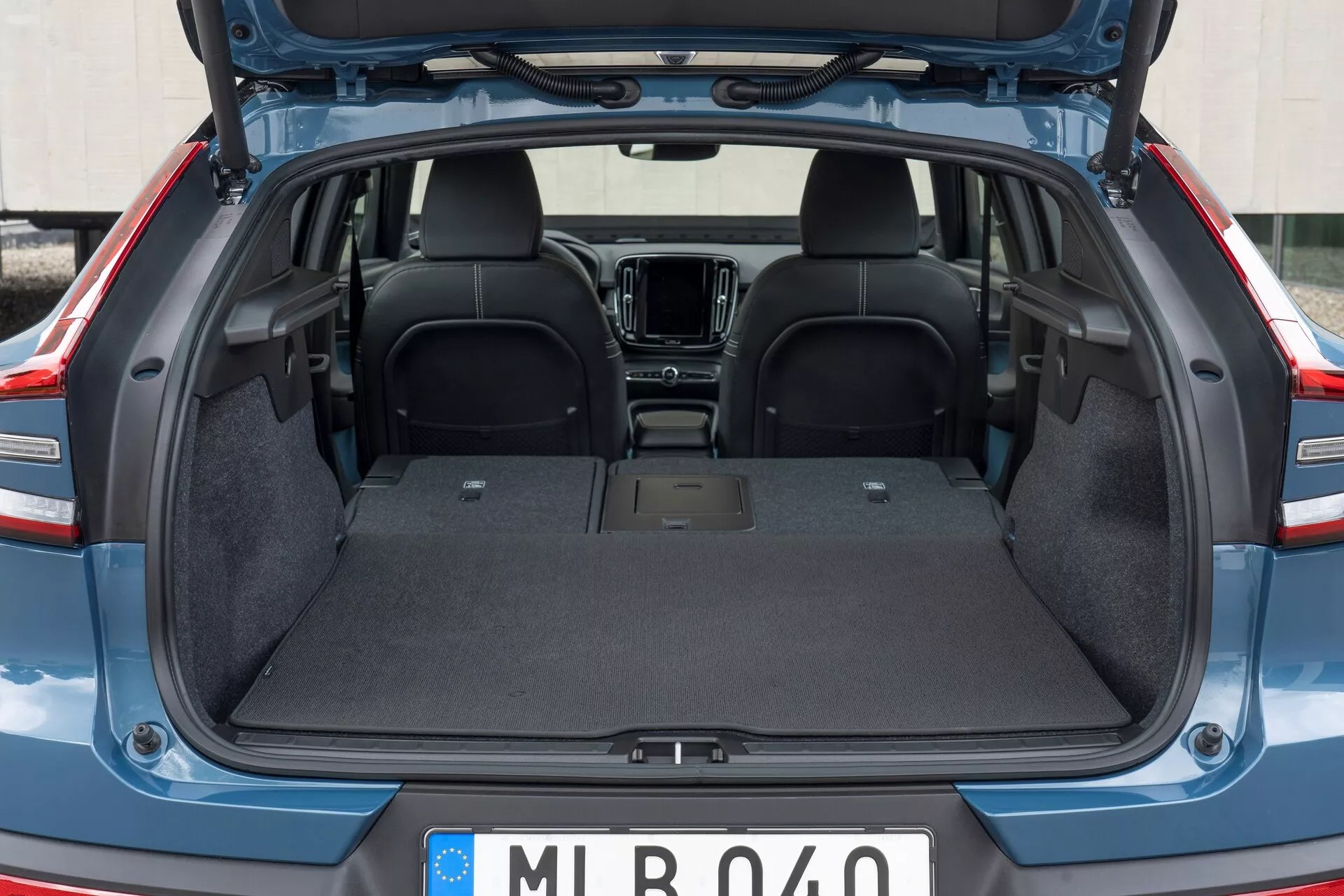 Wnętrze tylne Volvo C40 2021 (1. generacji) z otwartym bagażnikiem, pokazujące pustą przestrzeń ładunkową z wytrzymałą wykładziną dywanową, płaską powierzchnią i bez widocznych przeszkód. Widoczna jest górna część tylnej kanapy z ciemnego materiału, z częściowym widokiem na elementy przedniej kabiny, takie jak konsola środkowa i system informacyjno-rozrywkowy. Klapa bagażnika jest podniesiona, odsłaniając krawędzie z wykończeniem z tworzywa sztucznego/kompozytu.
