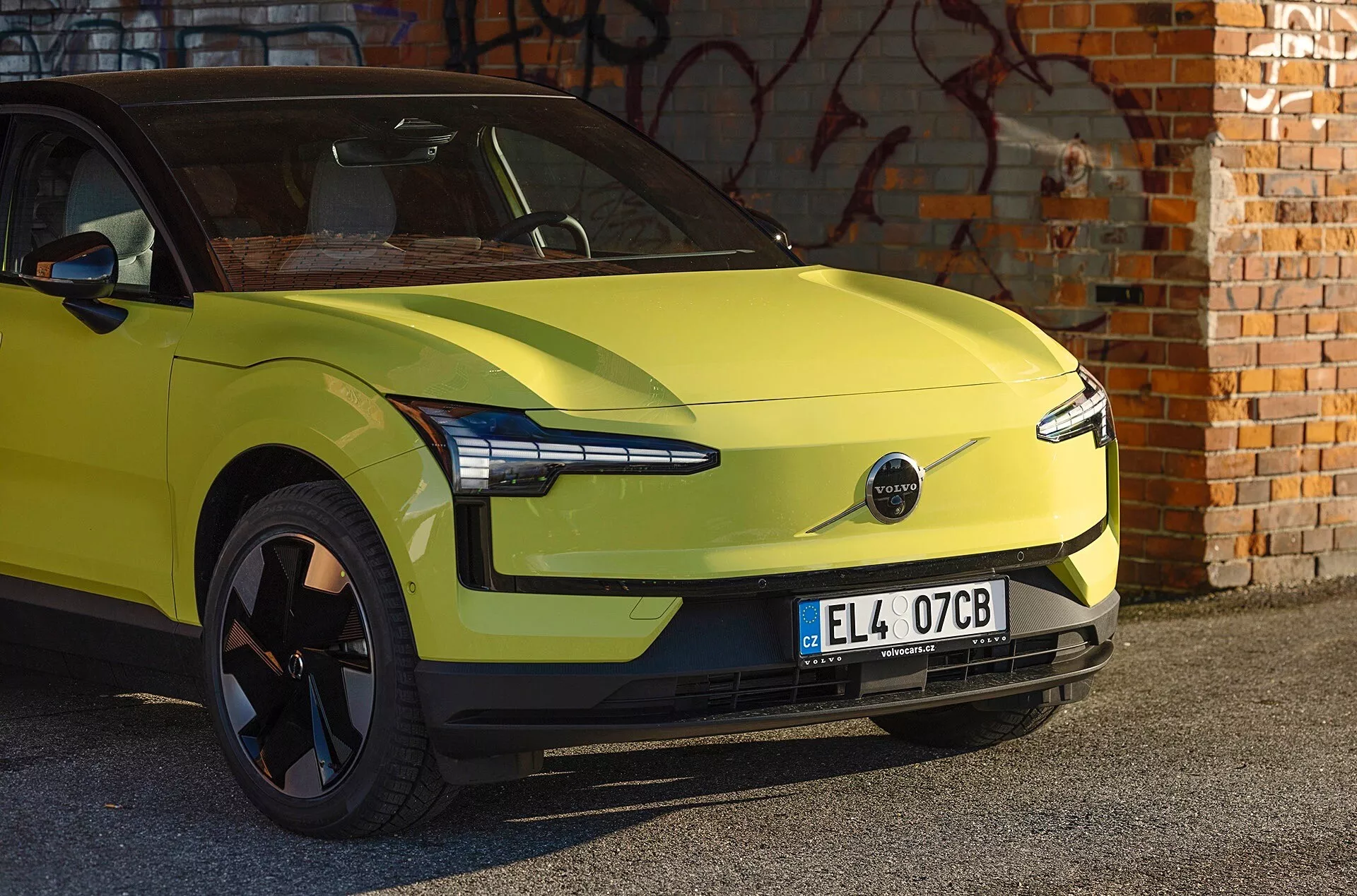 Limonkowo zielony samochód elektryczny Volvo EX30 z 2024 roku zaparkowany przed ścianą z cegły z graffiti, prezentujący aerodynamiczny design, nowoczesne reflektory LED i brak przedniej kratki