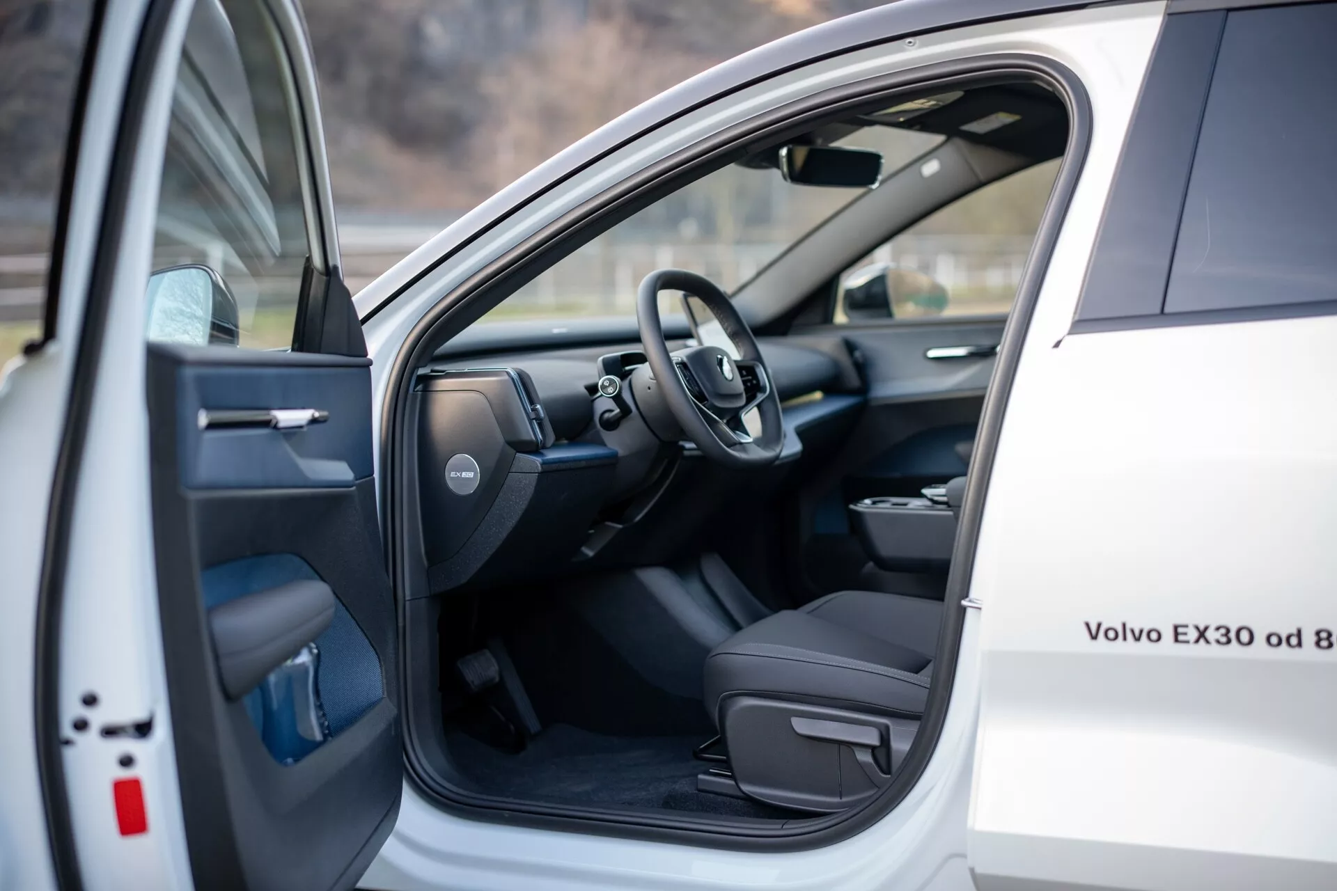 Widok wnętrza białego Volvo EX30 2024, pokazujący fotel kierowcy i obszar kokpitu z otwartymi przednimi lewymi drzwiami z elektrycznymi sterowaniami, oraz widok kierownicy i części deski rozdzielczej