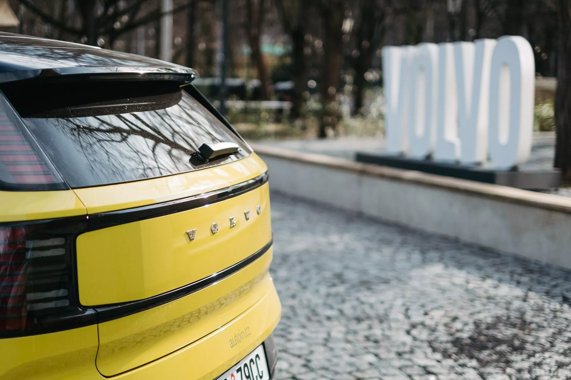 Zbliżenie na tylną część jaskrawożółtego Volvo EX30 2024, z poziomymi LEDowymi światłami tylnymi, potencjalną kamerą lub czujnikami z tyłu, pojedynczą wycieraczką tylnej szyby, oznaczeniem pojazdu elektrycznego lub hybrydowego i anteną w kształcie płetwy rekina, zaparkowanego na brukowanej powierzchni z rozmazanym znakiem 'OLVO' w tle