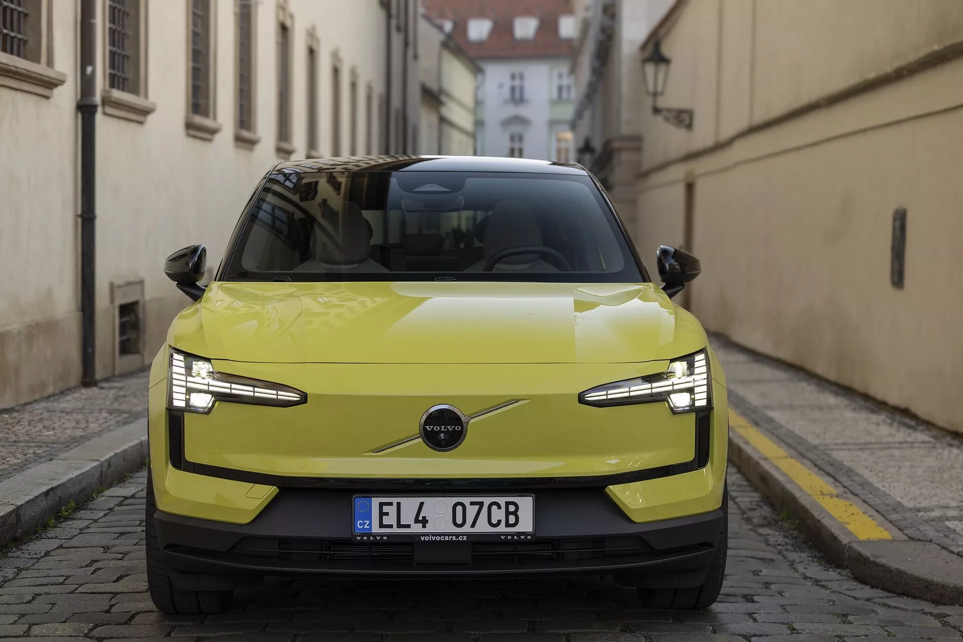 Przedni widok Volvo EX30 2024 w limonkowym żółtym kolorze, podkreślający jego eleganckie reflektory LED, minimalistyczną atrapę chłodnicy wskazującą na napęd elektryczny i zaawansowane funkcje technologiczne, takie jak czujniki parkowania i możliwe ADAS, na tle historycznej europejskiej ulicy