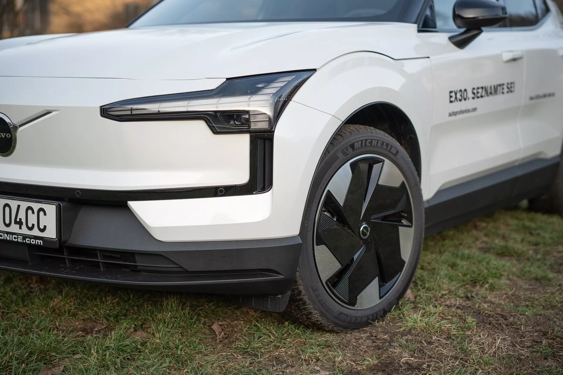 Zbliżenie na elektryczny pojazd Volvo EX30 2024 z perspektywy trzech czwartych, uwypuklające jego reflektor LED, aerodynamiczny design koła i minimalistyczny przedni zderzak bez kraty, w kolorze białym z czarnymi elementami.