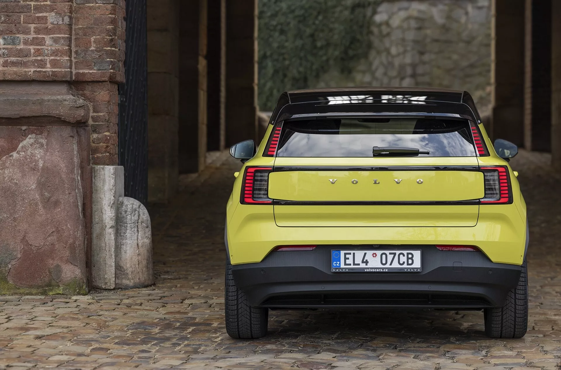 Tylny widok Volvo EX30 2024 na bruku ukazujący eleganckie poziome światła tylne, emblemat producenta, europejską tablicę rejestracyjną, dużą tylną szybę z rozmrażaczem, sportowy spojler i solidny zderzak z czujnikami parkowania