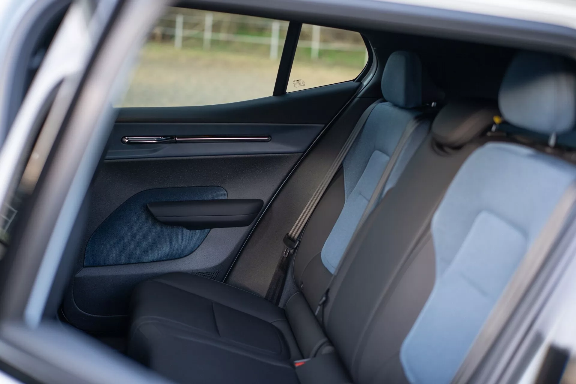 Wnętrze Volvo EX30 2024 przedstawiające tylną część pasażerską przez otwarte drzwi pasażera z przodu, cechujące się ciemną tapicerką, widocznymi zagłówkami, szczegółowym panelem drzwi z elementami sterującymi, projektem słupka B, pasami bezpieczeństwa na tylne siedzenia, częściowo widocznym bocznym oknem poprawiającym atmosferę wnętrza, oraz skupieniem na układzie siedzeń dla wygody i bezpieczeństwa pasażerów.