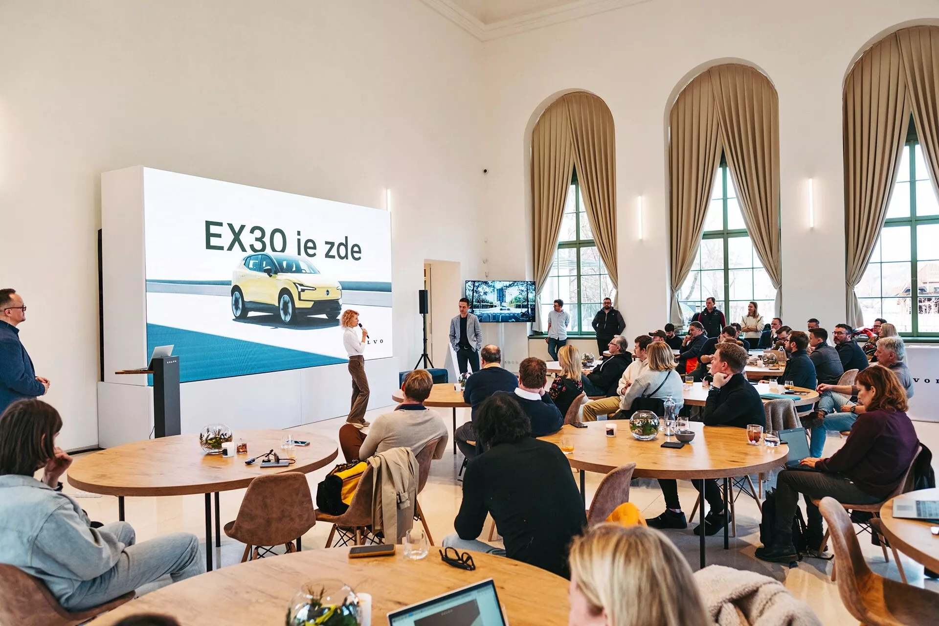 Prezentacja elektrycznego pojazdu Volvo EX30 2024 w przestronnej sali z wysokimi sufitami i wysokimi oknami łukowymi, przedstawiająca żółte EX30 z widoku trzech czwartych z przodu na dużym ekranie