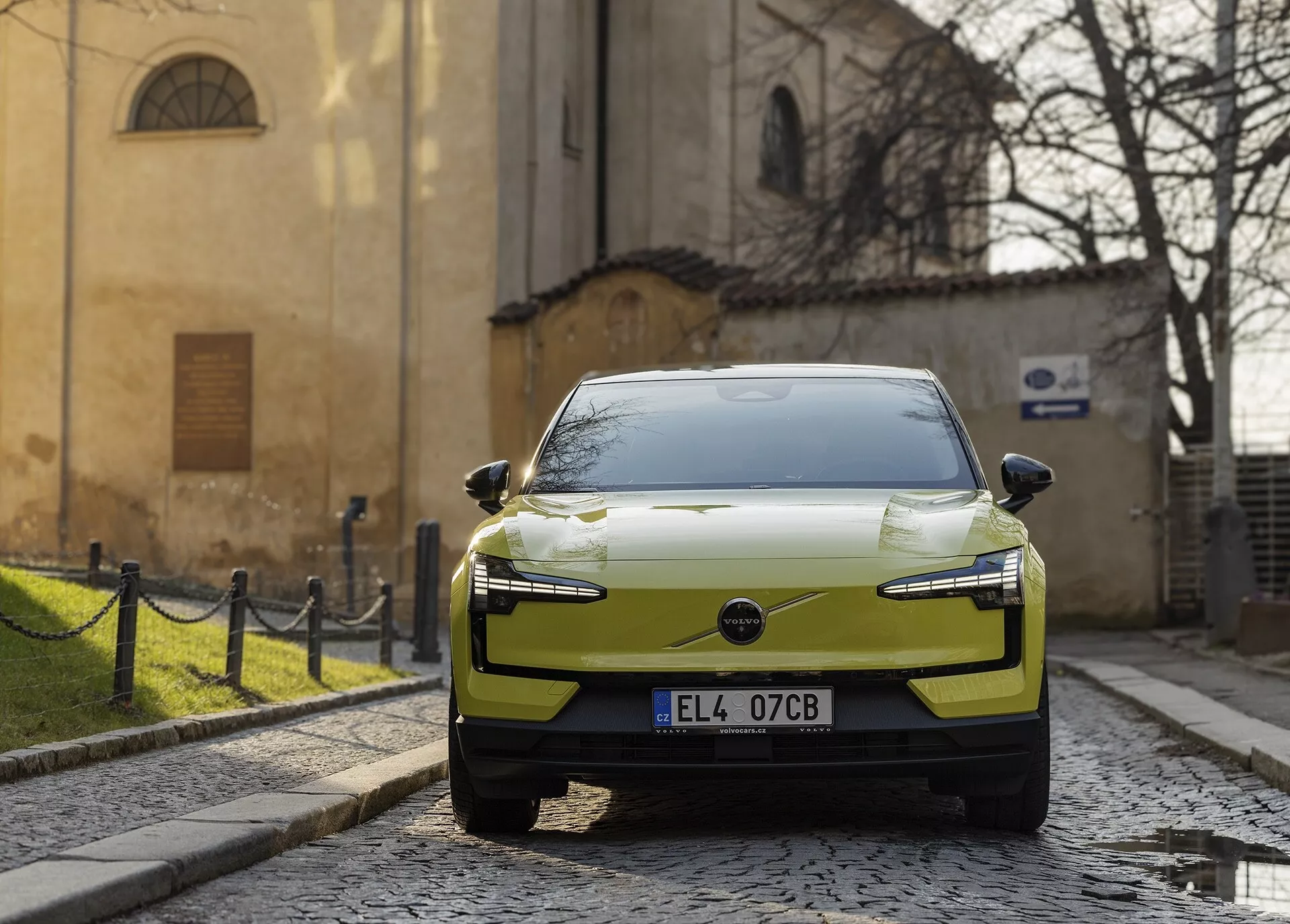 Nowoczesny, elektryczny samochód Volvo EX30 rok 2024 w kolorze jasnozielonym, zaparkowany na brukowanej ulicy, widziany z prawo-przedniej perspektywy z akcentowanymi diodowymi reflektorami i aerodynamicznym designem maski na tle historycznej europejskiej architektury.