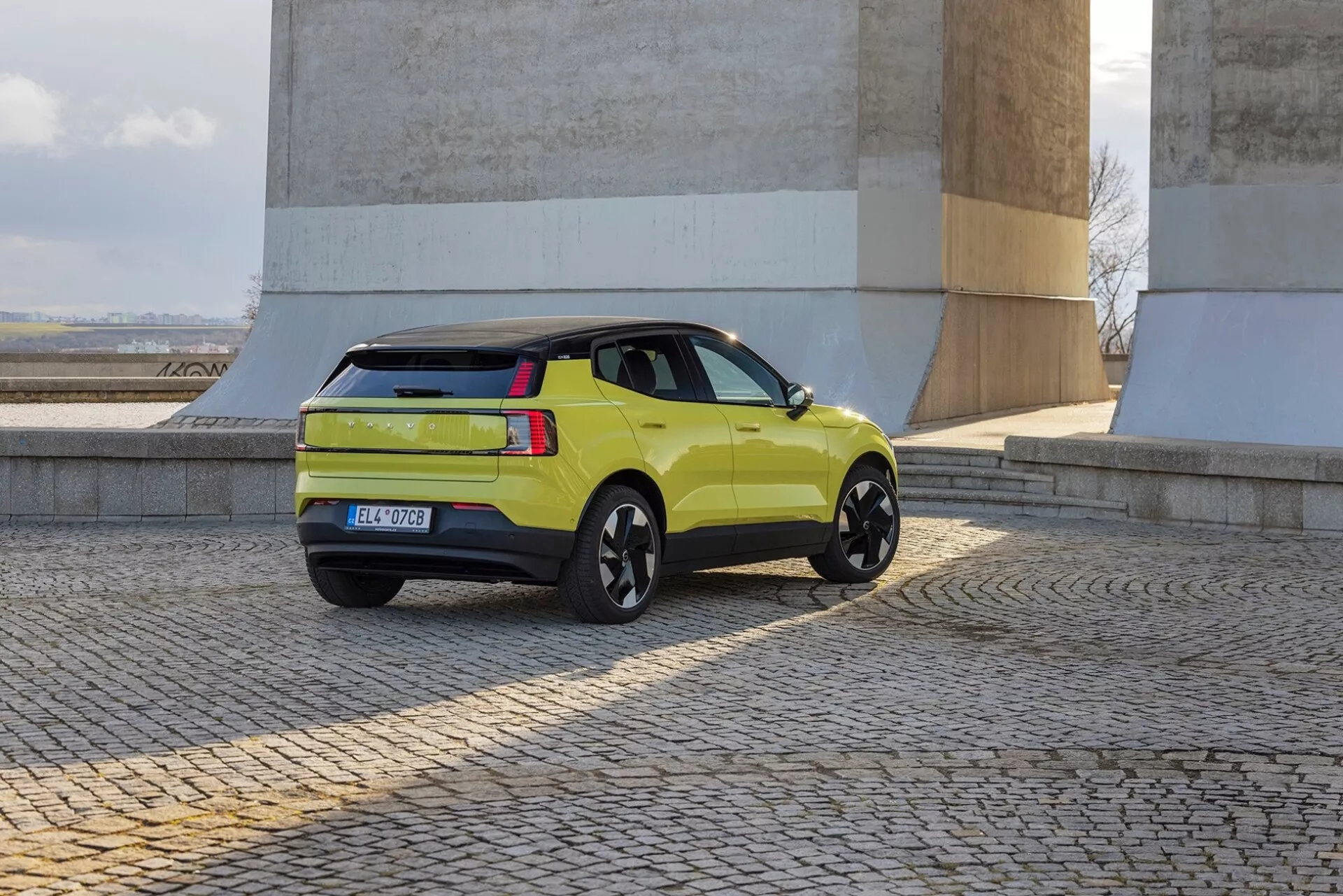 Limonkowe SUV Volvo EX30 2024 w widoku z tyłu z trzech czwartych ukazujące profil boczny, tylne światła, klapę bagażnika i czarne felgi aluminiowe w pobliżu kamiennych ścian na brukowanym podłożu