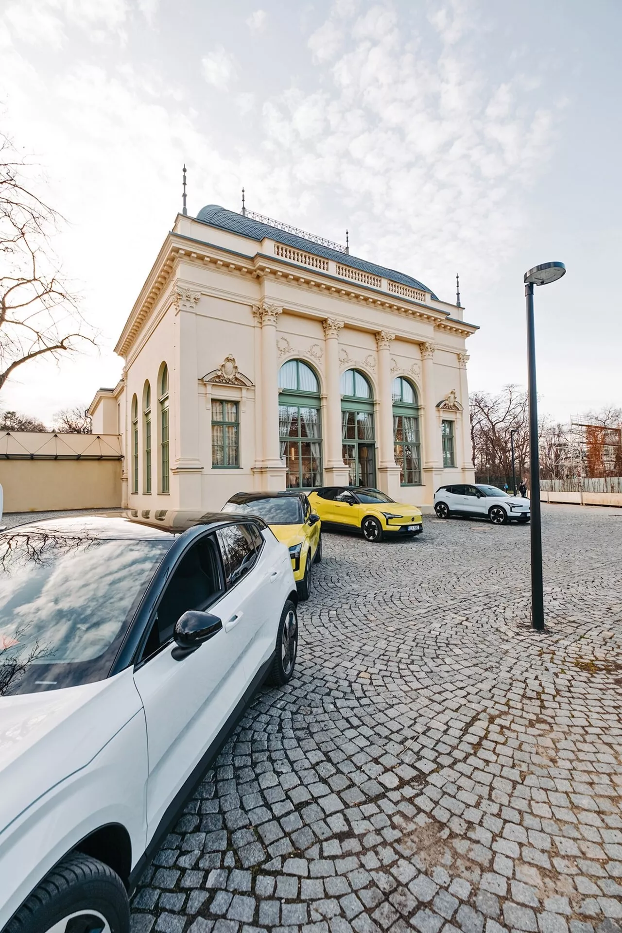 Scena uliczna z trzema samochodami i eleganckim budynkiem w tle, uchwycona z perspektywy szerokokątnego obiektywu ukazującego białą współczesną limuzynę, jaskrawożółty samochód sportowy i białą limuzynę lub hatchback w świetle późnego popołudnia