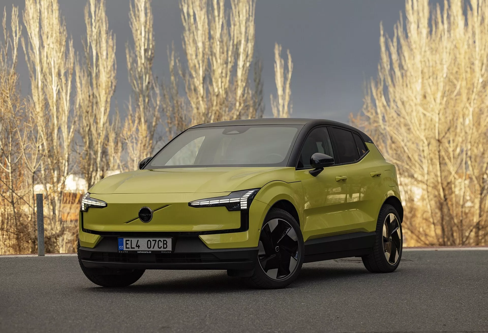 Nowoczesny limonkowo-zielony elektryczny samochód Volvo EX30 2024 w trzykwartalnym widoku z przodu, podkreślający jego eleganckie diody LED, brak tradycyjnej kratki dla lepszej aerodynamiki, duże czarne koła ze stopów, lusterka zewnętrzne w kolorze nadwozia z potencjalnie zintegrowaną technologią, gładki i aerodynamiczny profil boczny o wyglądzie zbliżonym do coupé, i żywy metaliczny wykończenie, zaparkowany w naturalnym otoczeniu