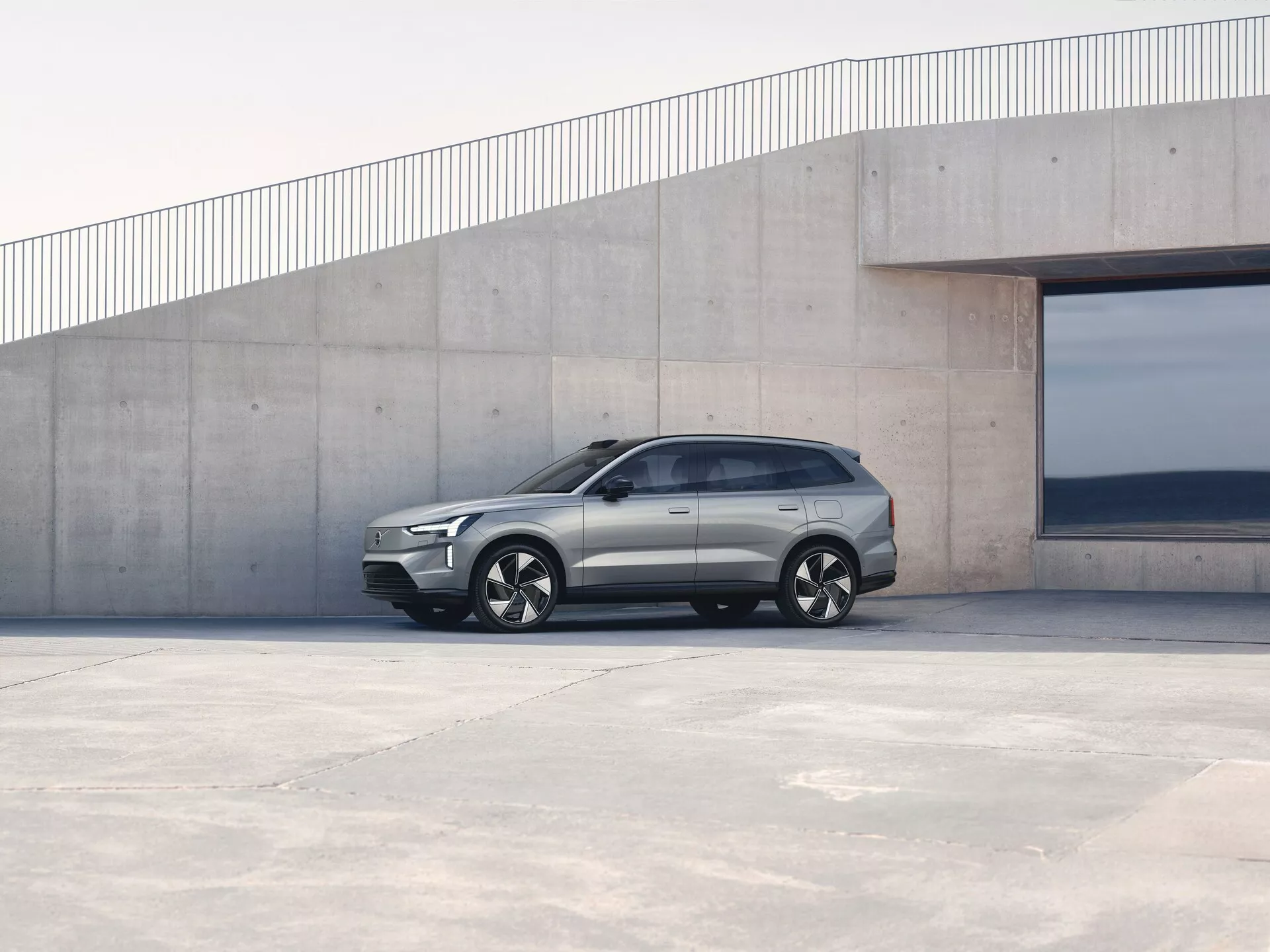 Volvo EX90 1. generacji z 2023 roku zaparkowane na zewnątrz, widok z profilu bocznego pokazujący kątowy design, klamki drzwiowe w linii z karoserią i felgi aluminiowe na tle minimalistycznej architektury