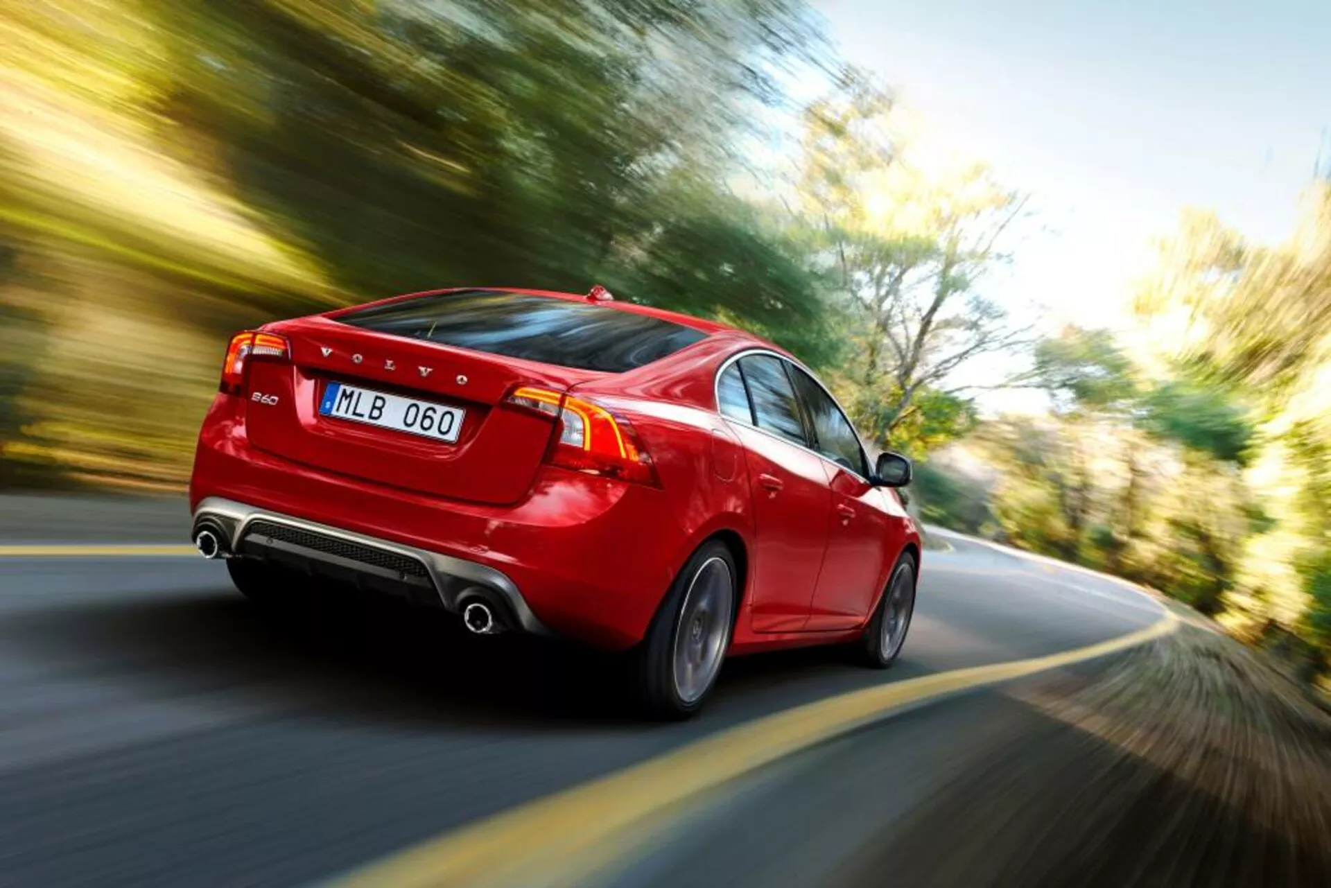Dynamiczny widok z tyłu i boku czerwonego sedana Volvo S60-V60 2004 w ruchu, pokazujący LED-owe tylne światła, podwójny wydech, stylowe felgi aluminiowe i wyraźne oznaki prędkości poprzez efekt rozmycia ruchu.