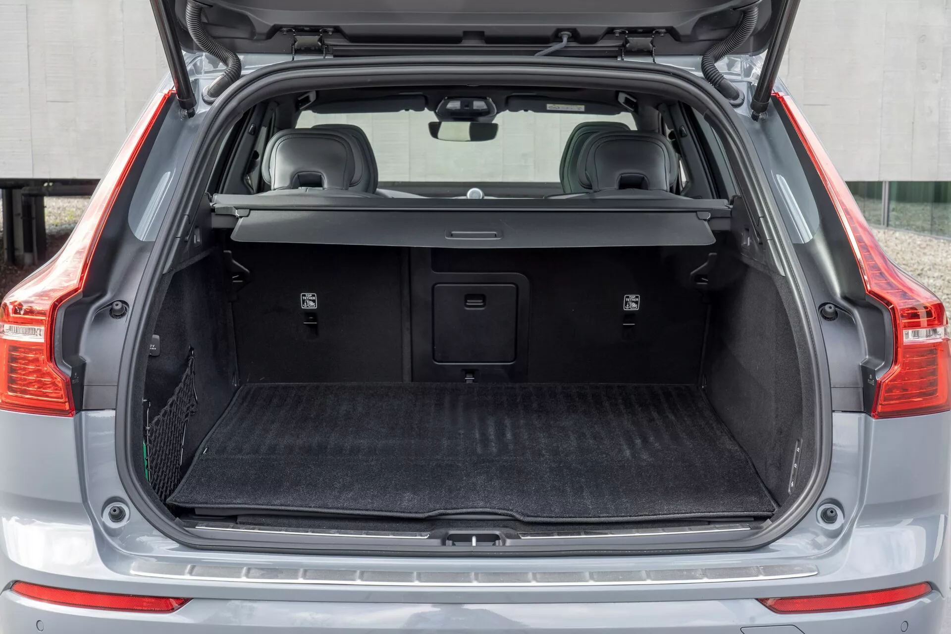 Tylny przedział ładunkowy Volvo XC60 2021 (druga generacja facelift), pokazujący otwarty bagażnik z prostego widoku tylnego z pustą przestrzenią ładunkową, częściowo odsuniętą zasłoną bagażową, matą ochronną, plastikowymi wykończeniami z małymi schowkami, możliwym gniazdem 12V, metalową nakładką progu i składanymi tylnymi siedzeniami z zagłówkami.