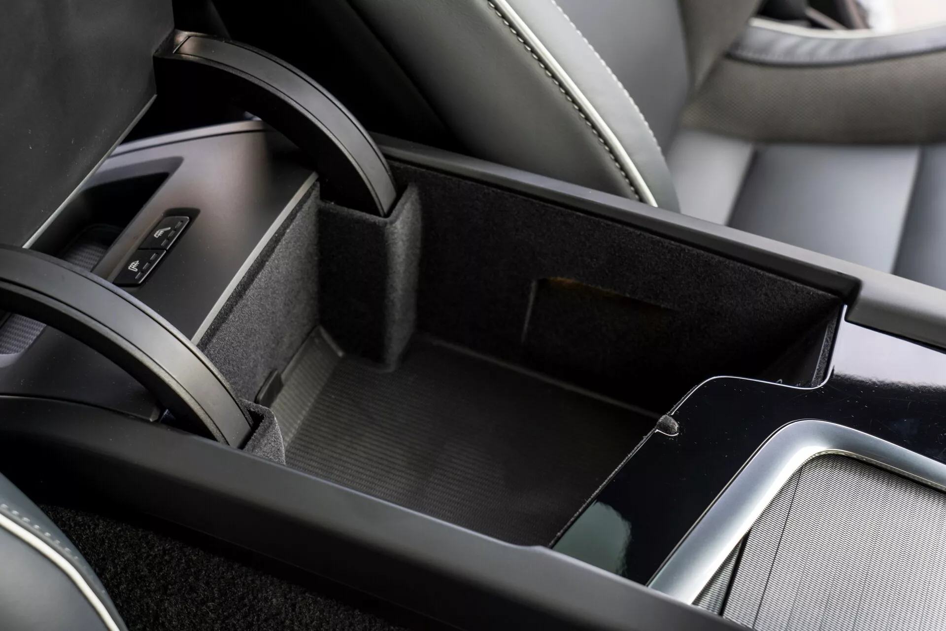 Zbliżenie obszaru konsoli środkowej w wnętrzu Volvo XC60 2021, ukazujące otwarty schowek wyściełany materiałem przypominającym filc, dwupoziomowe przechowywanie z mniejszą górną tacką, skórzany podłokietnik i metaliczne wykończenia dodające elegancji. Na krawędzi obrazu widoczna jest część obszaru dźwigni zmiany biegów z osłoną ze skóry.