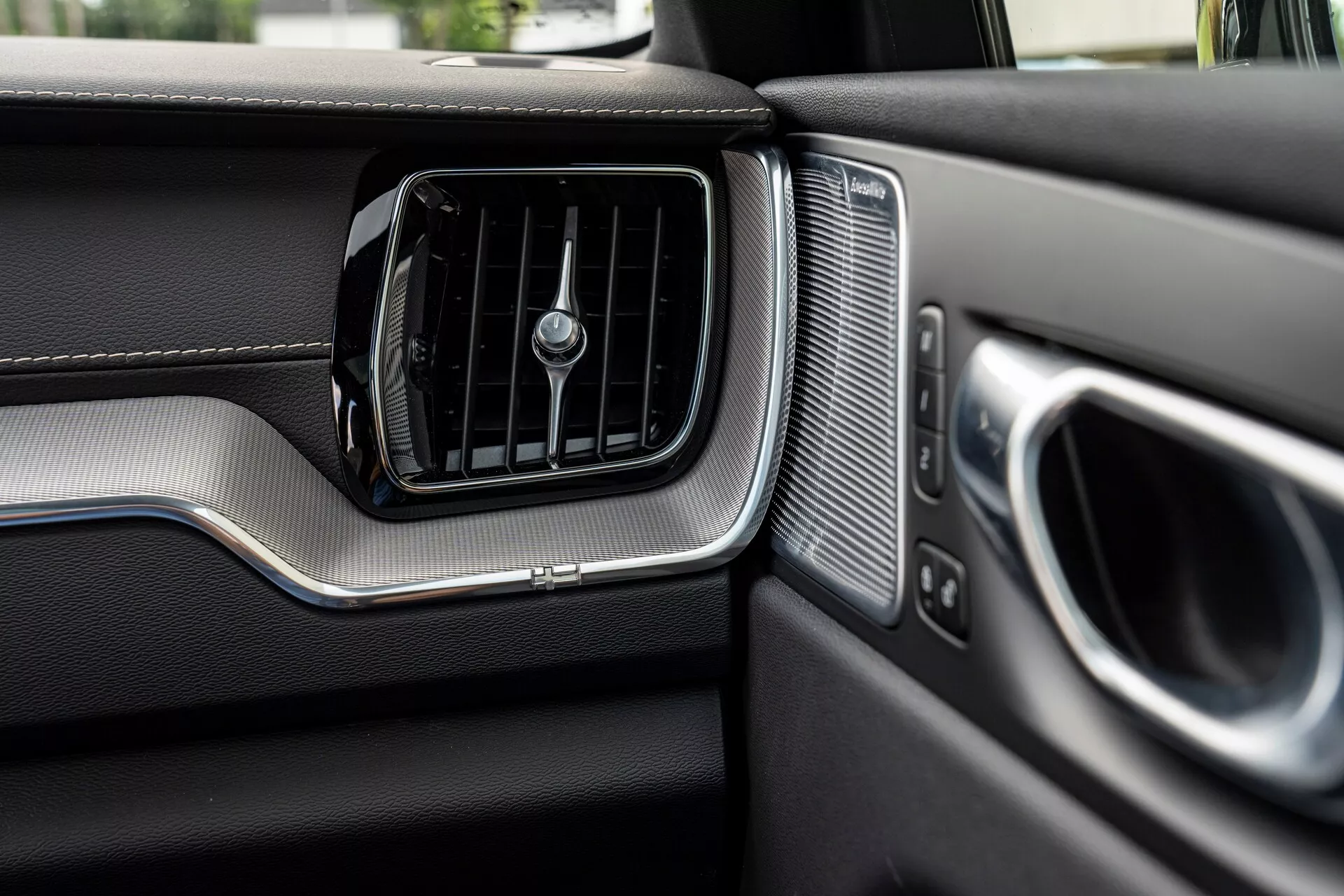 Zbliżenie deski rozdzielczej po stronie pasażera w Volvo XC60 2021 (2. generacja, lifting). Obraz wyraźnie pokazuje kratkę wentylacji klimatyzacji z poziomymi żaluzjami oraz centralnym obrotowym pokrętłem, otoczoną szczotkowanym metalowym akcentem. Po prawej stronie kratki znajduje się grill głośnika z równomiernymi perforacjami, obok częściowo widocznego podłokietnika i klamki drzwi, również z metalowymi akcentami. Deska rozdzielcza prezentuje materiały podobne do skóry w górnej części i inaczej teksturowany dolny obszar oddzielony metalowym paskiem, co podkreśla premium design i jakość wykonania pojazdu.
