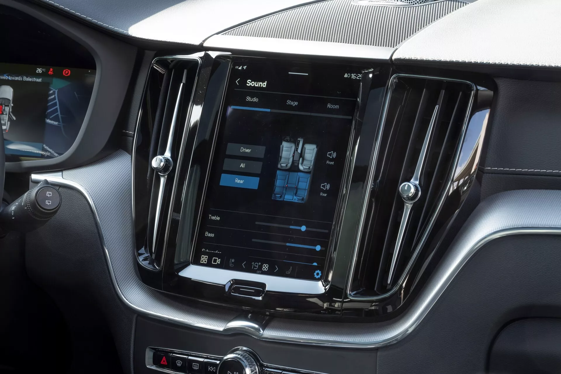 Zdjęcie wnętrza Volvo XC60 2021 facelift, skupiające się na konsoli środkowej z dużym pionowym ekranem dotykowym systemu infotainment. Zrobione z perspektywy siedzenia pasażera, obraz pokazuje również ustawienia dwóch stref klimatyzacji, metalowe wloty wentylacyjne oraz przyciski manualne pod ekranem, co nadaje nowoczesny i luksusowy wygląd.