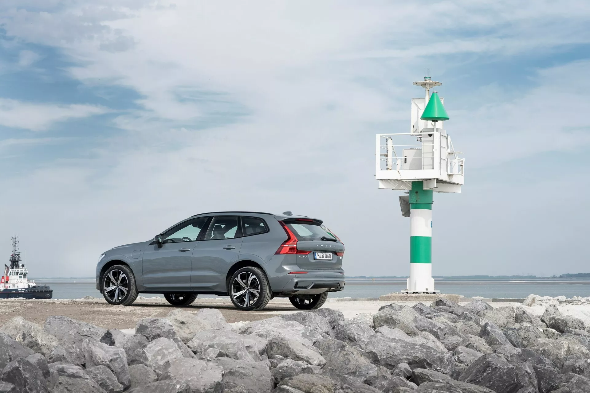 Fotografia modelu Volvo XC60 2021 drugiej generacji po liftingu, uchwycona pod kątem trzech czwartych z tyłu na skalistej powierzchni, z morskim tłem w tle. Ciemnoszary samochód z metalicznym wykończeniem posiada elegancką, sportową linię dachu, aluminiowe felgi wieloramienne, tylne czujniki parkowania i podwójny wydech. Po prawej stronie samochodu widoczna jest mała latarnia morska lub sygnał nawigacyjny, a w wodzie holownik lub pilotówka, co podkreśla nadmorski charakter.