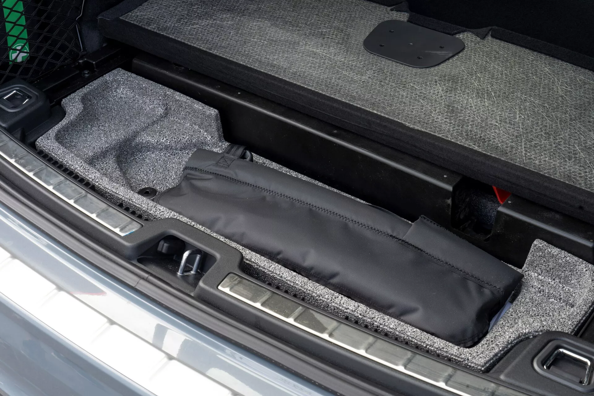 Zbliżenie na obszar ładunkowy Volvo XC60 2021 z ukrytym schowkiem pod podłogą bagażnika z czarną torbą, wyściółką z dywanu i widocznym metalowym progiem, obok czerwonego narzędzia awaryjnego.
