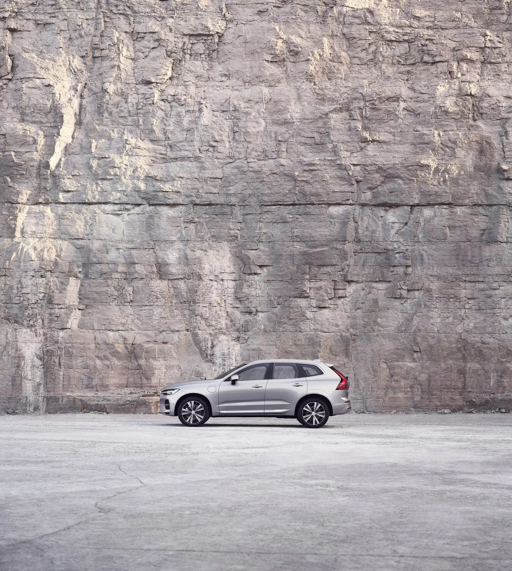 Widok z profilu bocznego na Volvo XC60 2021 (2. generacja facelift) zaparkowany przed dużą teksturowaną ścianą skalną lub betonową, prezentujący jego elegancki i luksusowy design z ciemną metaliczną farbą, wieloramiennymi felgami, przyciemnionymi szybami i czarnymi słupkami B w miękkim, rozproszonym świetle.