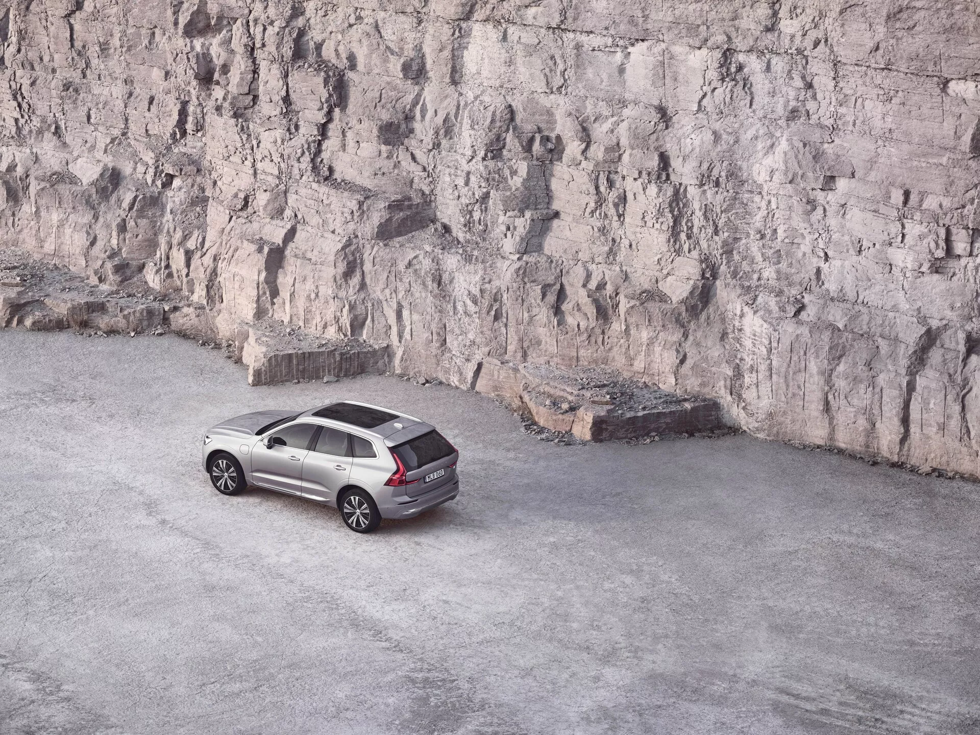 Srebrny Volvo XC60 2021 drugiej generacji facelift zaparkowany na otwartym, żwirowym terenie z dużym skalnym urwiskiem w tle, widziany z podwyższonego kąta 45 stopni, podkreślający górny i boczny profil eleganckiego crossover SUV. Godne uwagi cechy to duże obręcze ze stopu wieloramiennego, charakterystyczne reflektory i tylne światła LED, relingi dachowe i aerodynamiczne linie projektowe.