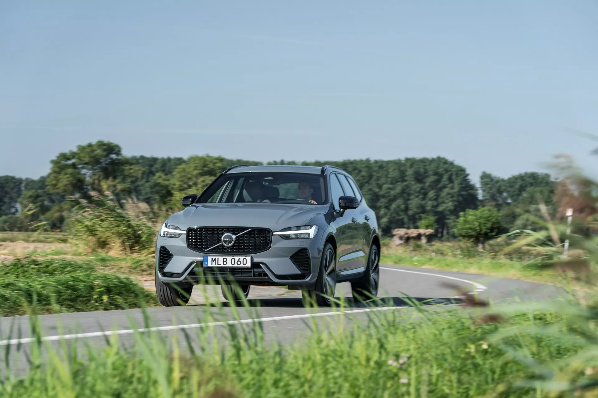 Nowoczesny SUV, Volvo XC60 2021 (facelifting 2. generacji), uchwycony pod niskim kątem trzy czwarte z przodu, podkreślający dynamiczny wygląd i cechy designu. Wyrazista kratka z dużym emblematem, eleganckie reflektory LED i rzeźbiony zderzak z wbudowanymi wlotami powietrza. Aerodynamiczny, luksusowy wygląd z wyprofilowanymi liniami, wysokim prześwitem, dużymi wieloramiennymi felgami aluminiowymi, oponami niskoprofilowymi, lusterkami bocznymi z wbudowanymi kierunkowskazami, pochylonym dachem i przyciemnianymi szybami. Sugeruje zaawansowane technologie i funkcje bezpieczeństwa, jazda po wiejskiej drodze z rozmazaną roślinnością sugerującą ruch.