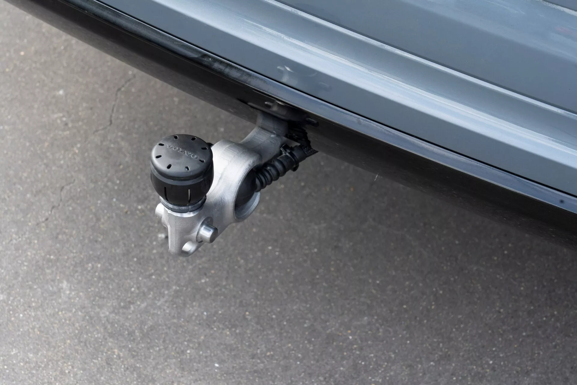 Zbliżenie wysuwanego stopnia bocznego w Volvo XC60 2021 (2-gie pokolenie-lifting), przedstawiające rozszerzoną powierzchnię antypoślizgową z wypukłymi czarnymi kropkami pod drzwiczkami bocznymi pojazdu, przyczepioną do mechanizmu zawiasowego z metalu. Fotografowane z niskiego kąta, podkreśla zautomatyzowaną funkcję zaprojektowaną, aby ułatwić wsiadanie i wysiadanie przez zmniejszenie odległości od ziemi.