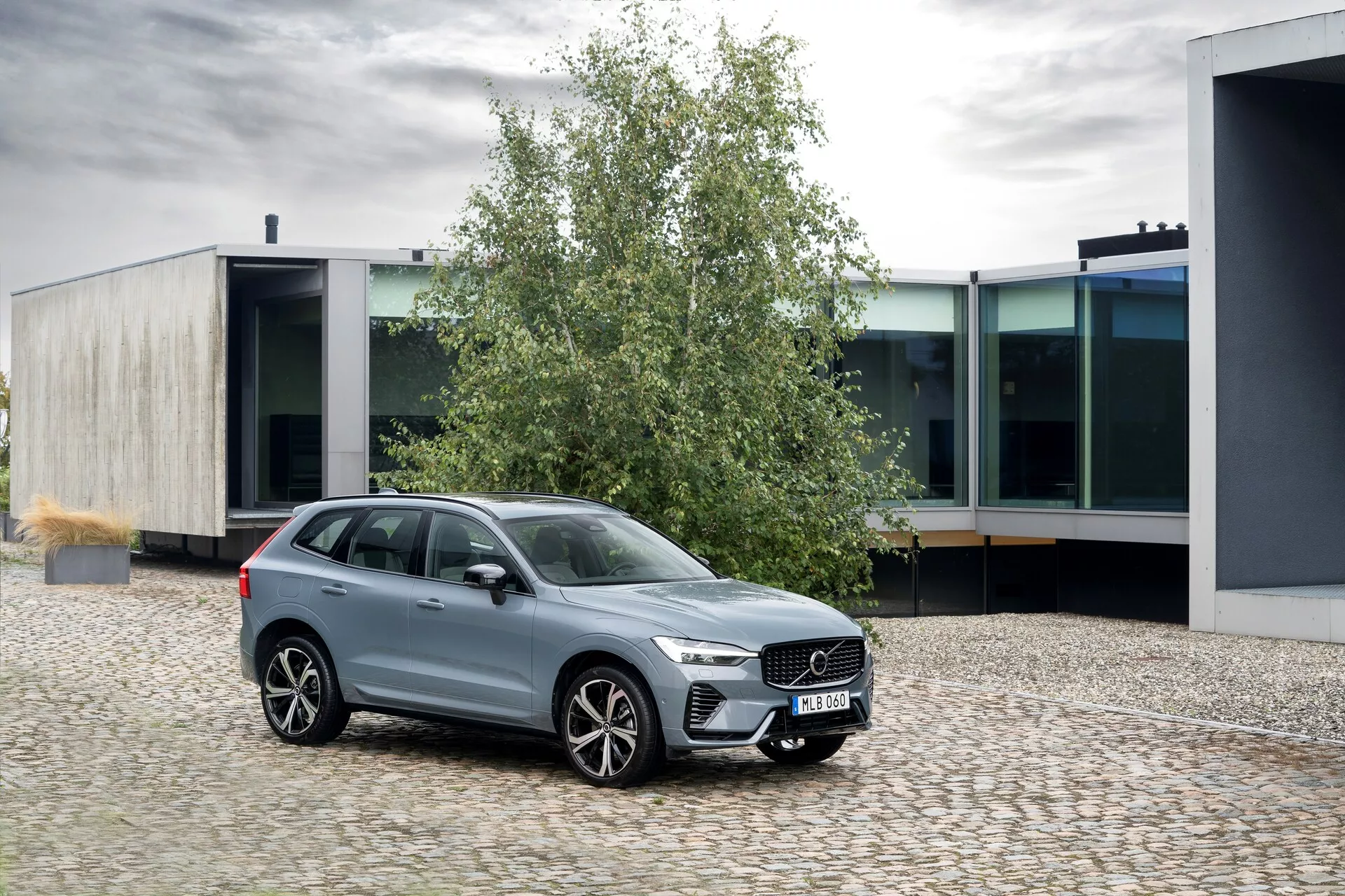 Obraz Volvo XC60 2021 (druga generacja - lift) zaparkowanego na brukowanej powierzchni, ustawionego pod lekkim kątem w lewą stronę. Widok z przodu z trzema czwartego podkreśla wyrazistą kratkę z pionowymi prętami, smukłe reflektory LED z dziennymi światłami oraz duże wieloszprychowe felgi aluminiowe. Okna są otoczone chromem, co wzmacnia jego polerowane metaliczne wykończenie. SUV ma podwyższony prześwit, relingi dachowe i lusterka boczne w kolorze nadwozia z zintegrowanymi kierunkowskazami. Sceneria obejmuje środowisko miejskie z nowoczesną architekturą, ukazując elegancki design i luksusowe cechy pojazdu.