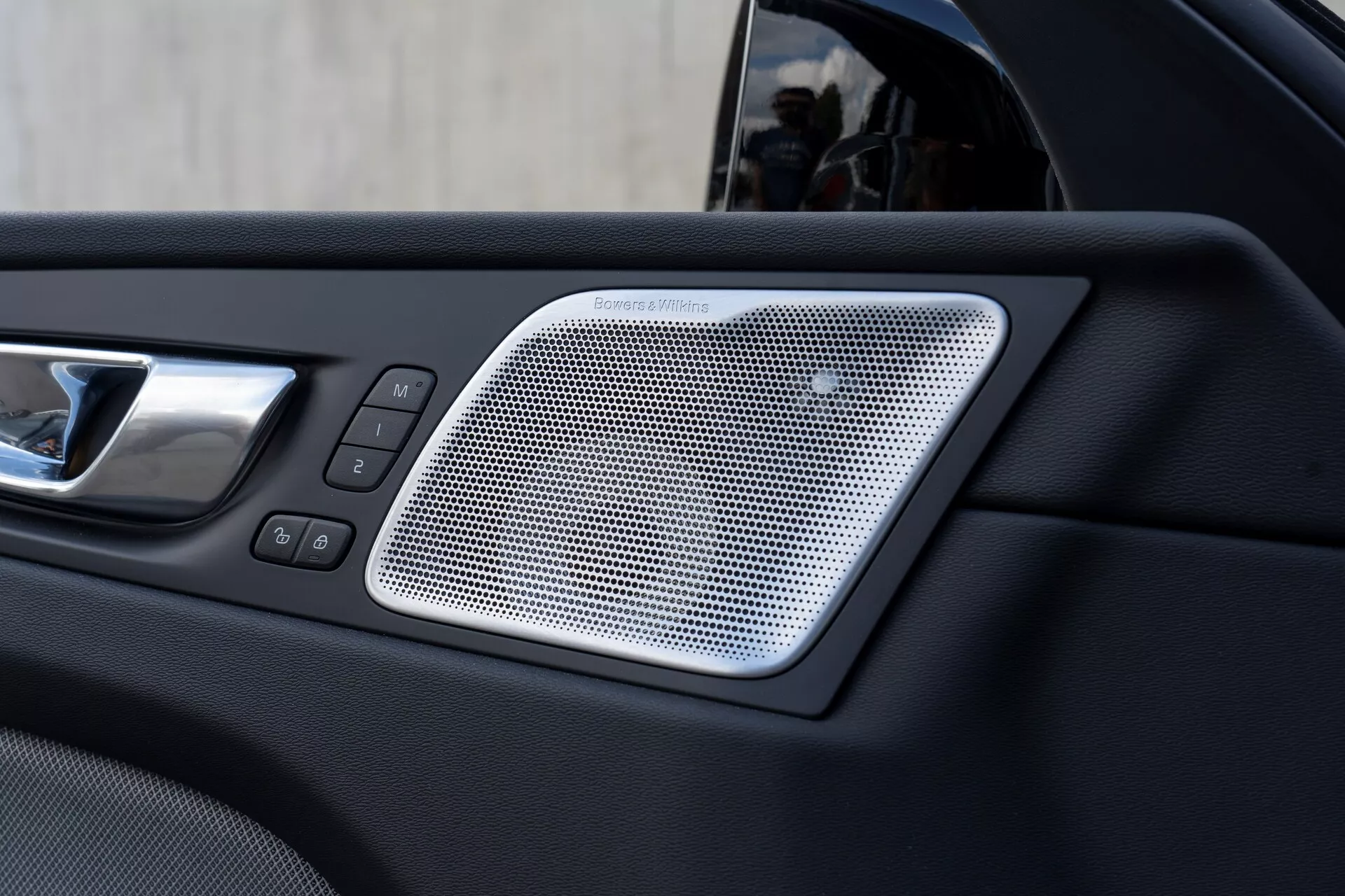 Zbliżenie wnętrza drzwi samochodu Volvo XC60 2021 (2. generacja, lifting) ukazujące kratkę głośnika Harman/Kardon o siatkowym wzorze, chromowaną klamkę drzwi i sterowanie oknami oraz zamkami elektrycznymi, podkreślając luksus i nowoczesność funkcji pojazdu.