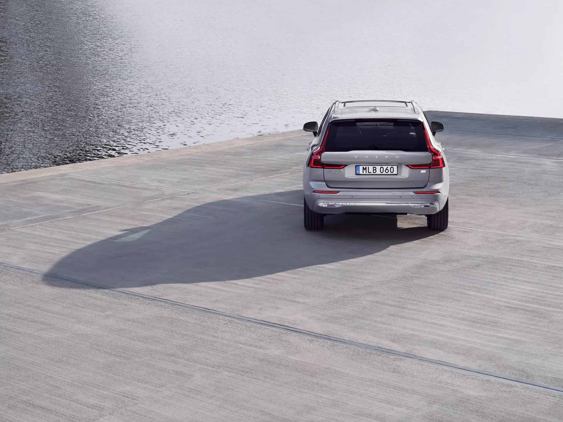 Widok z tyłu srebrnego SUV-a Volvo XC60 2021 (druga generacja, lifting) zaparkowanego na betonowej powierzchni w pobliżu zbiornika wodnego, z widocznym spojlerem dachowym, tylnymi światłami LED, klapą bagażnika, tylnym zderzakiem i rurami wydechowymi z wysokiego kąta z rzucanymi cieniami, w spokojnych warunkach przy lekkim wietrze.