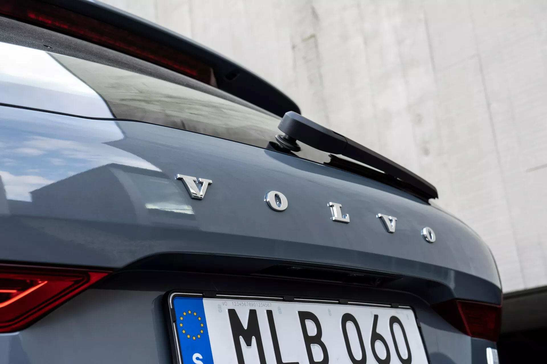 Zbliżenie tylnej części samochodu Volvo XC60 2021 (druga generacja facelift). Na obrazie widać wyróżniające się srebrne litery tworzące napis 'Volvo' na tylnym hatchbacku, część tylnej szyby i zintegrowany tylny spoiler z trzecim światłem stop LED. Samochód jest ciemnoszary lub grafitowy, z pionowym tylnym światłem LED po lewej stronie, który ma wzór ‘młota Thora’. Pod nazwą marki znajduje się tablica rejestracyjna w europejskim stylu z flagą UE, co wskazuje na rejestrację w UE, oprawiona chromowaną ramką. Kąt kamery jest lekko przesunięty w prawo, skupiając się na literach i krawędziach nadwozia samochodu, z neutralnym białym tłem.