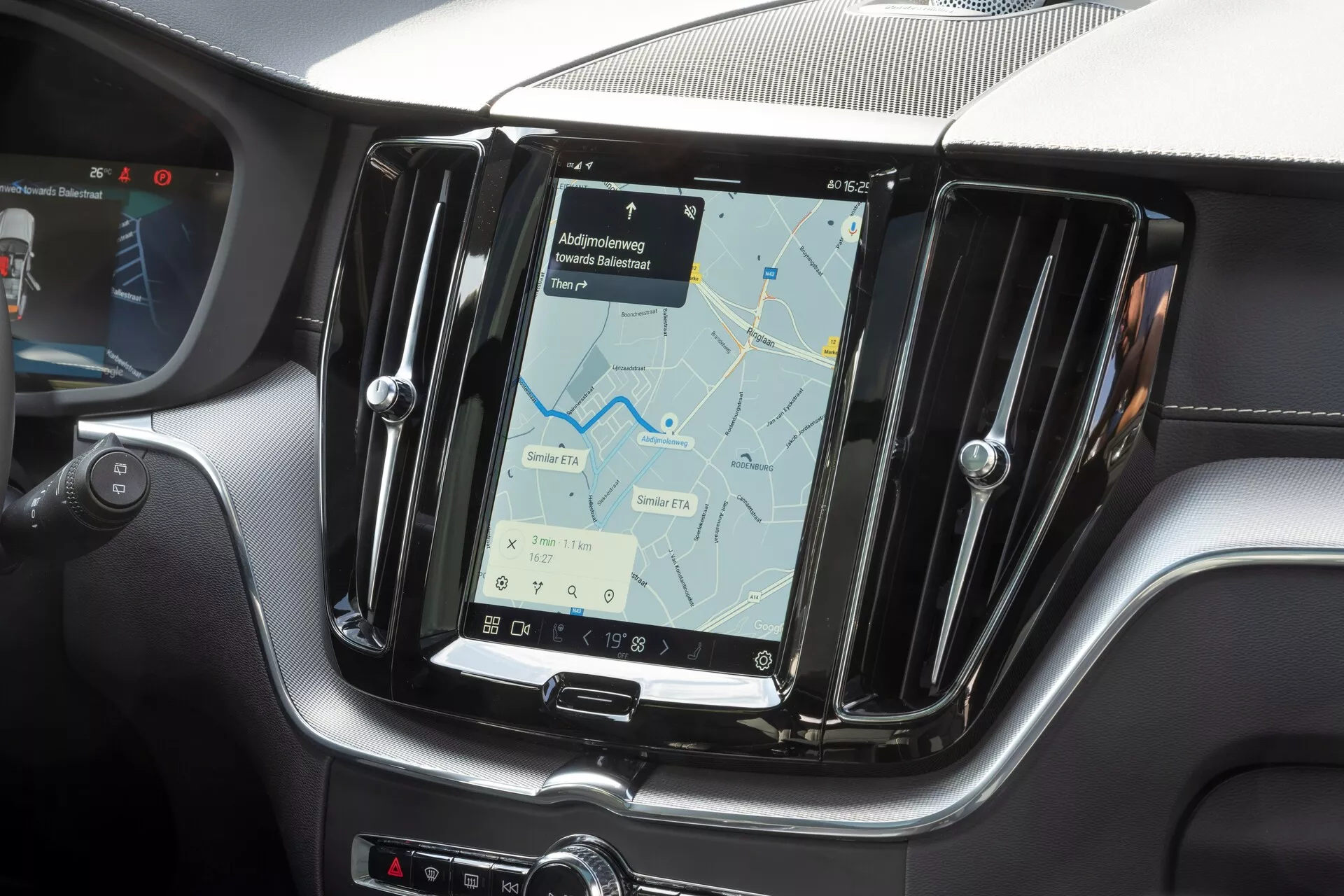 Zbliżenie konsoli środkowej w Volvo XC60 2021 (2. generacja, lifting), skupiając się na systemie infotainment. Zdjęcie, wykonane z perspektywy pasażera z przodu, pokazuje duży pionowy ekran dotykowy wyświetlający mapę nawigacyjną. Ekran jest otoczony metaliczną ramką i otoczony teksturą przypominającą włókno węglowe, podkreślając nowoczesne i sportowe elementy wnętrza. Fizyczne przyciski są widoczne na boku ekranu i poniżej w celu kontroli klimatu, co wskazuje na połączenie funkcji dotykowych i fizycznych. Częściowy widok kierownicy na lewym krańcu potwierdza konfigurację z lewostronnym napędem.