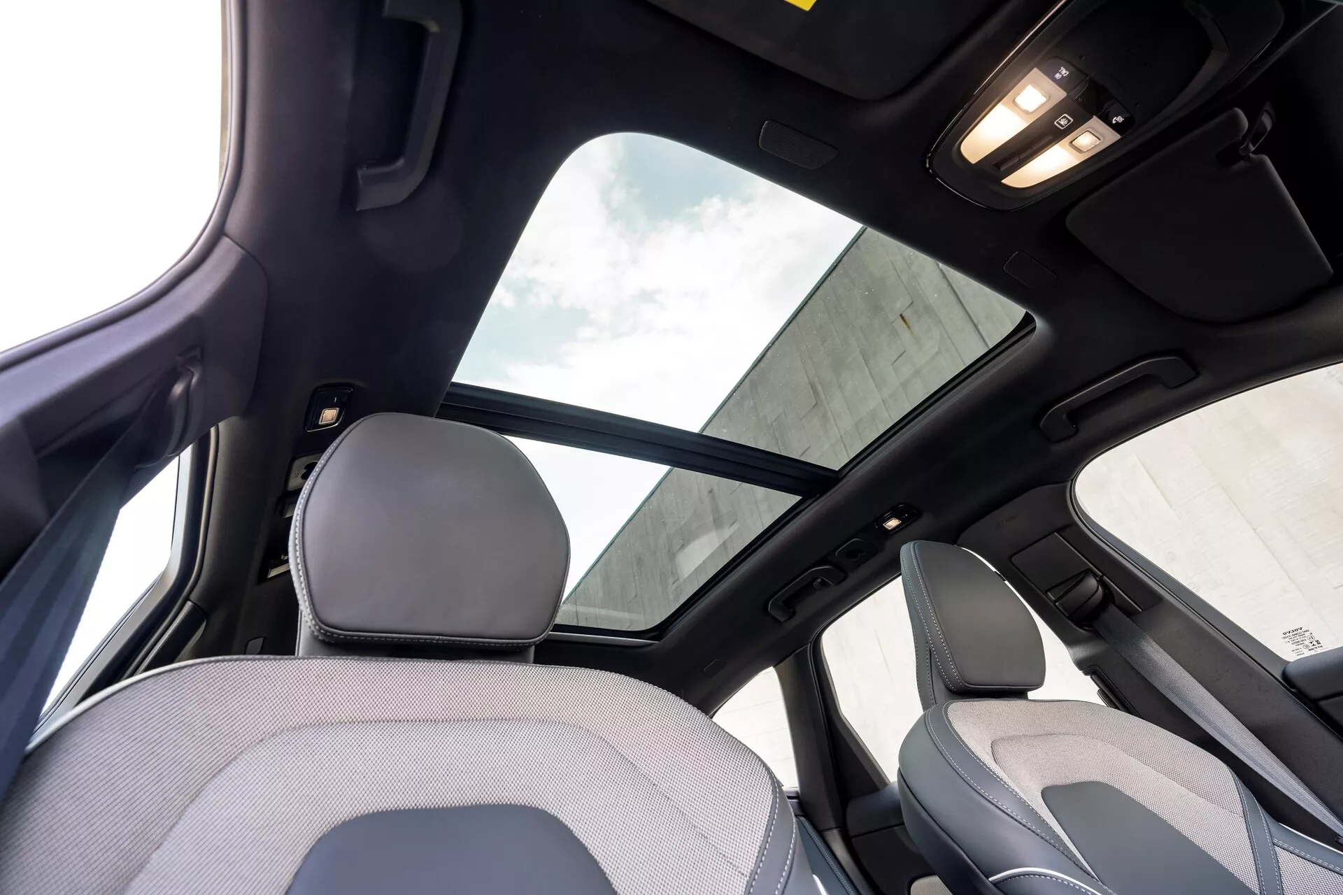 Wnętrze Volvo XC60 2021 2. generacji po liftingu widziane z tylnego siedzenia, koncentrujące się na górnej części kabiny. Charakterystyczne cechy obejmują duże panoramiczne okno dachowe z osłoną przeciwsłoneczną, czarną podsufitkę wskazującą na wysoki poziom wyposażenia lub pakiet luksusowy oraz oświetlenie wnętrza na suficie. Ujęcie ukazuje część zagłówka i górnej części oparcia przedniego siedzenia pasażera, a także słupek B i uchwyt nad tylnymi drzwiami.