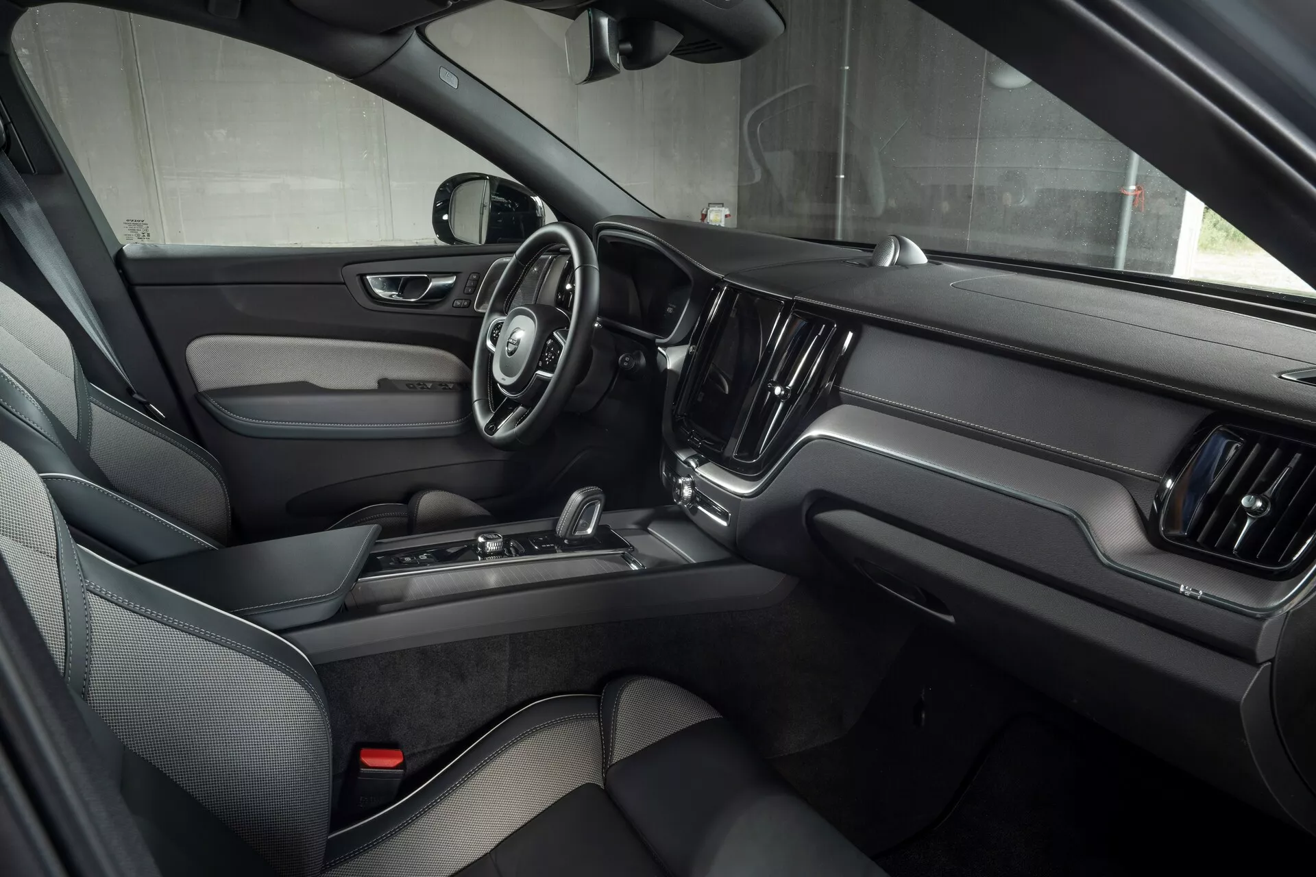 Widok wnętrza Volvo XC60 2021 (lifting 2. generacji) z perspektywy przedniego siedzenia pasażera, podkreślający drzwi kierowcy z elektronicznymi systemami sterowania, trójramienną kierownicę z wielofunkcyjnymi przyciskami, duży centralny ekran dotykowy systemu infotainment oraz luksusowy design z ciemną skórą i metalicznymi akcentami.