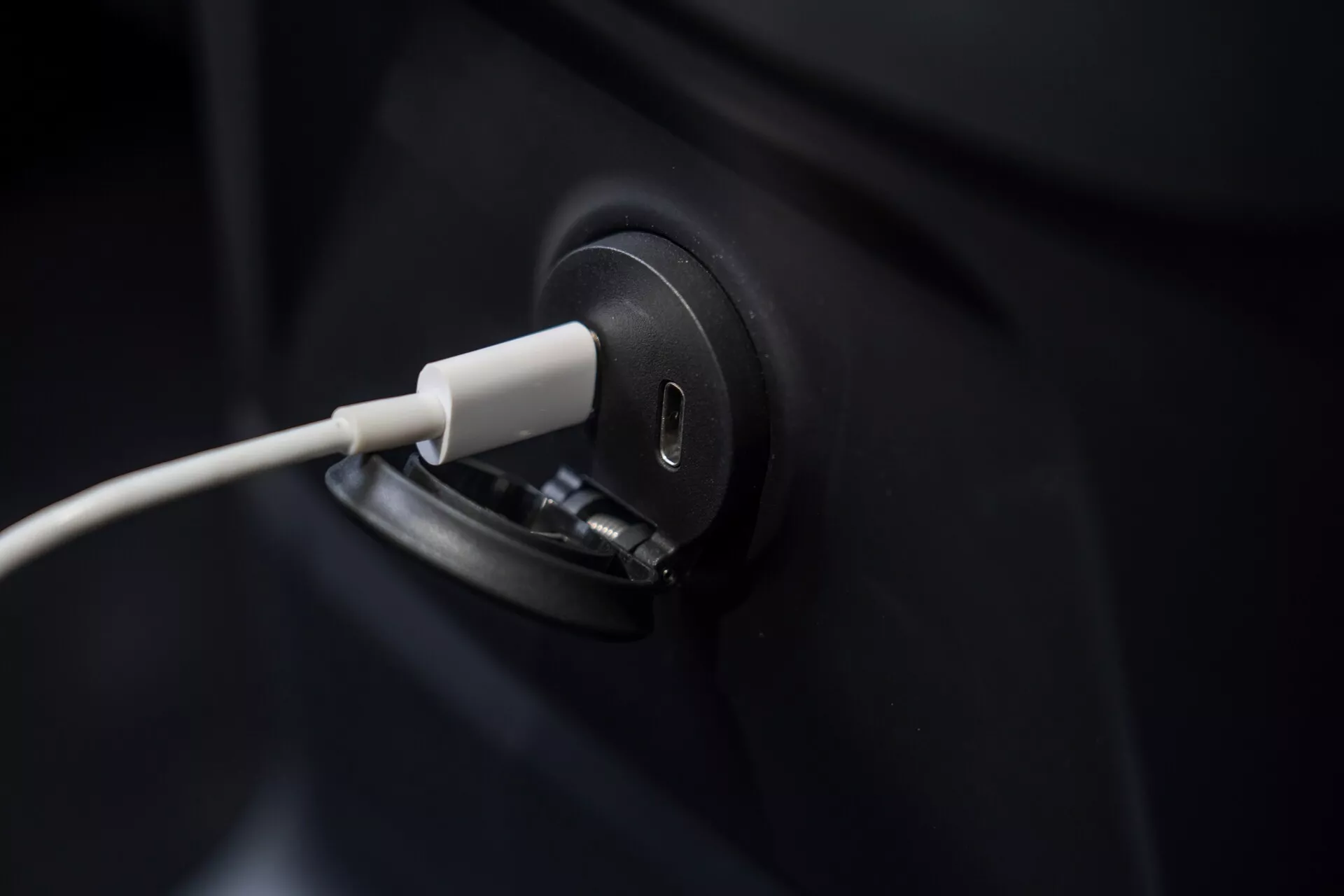 Zbliżenie na port ładowania elektrycznego pojazdu Volvo XC60 2021 z podłączonym kablem, podkreślając smukły i nowoczesny design. Kadrowanie skoncentrowane na porcie ładowania, pozostawiając ciemne nadwozie pojazdu w dużej mierze niewidoczne. Rozmyte tło podkreśla precyzję procesu ładowania pojazdu elektrycznego, symbolizując zieloną technologię i zrównoważony transport.