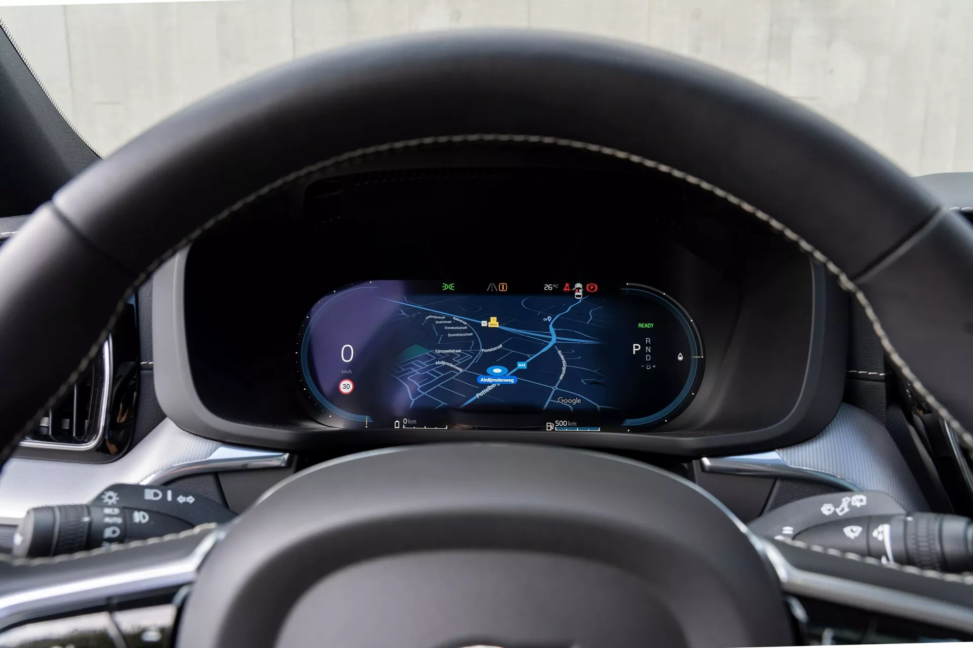 Zbliżenie na deskę rozdzielczą Volvo XC60 2021 (2. generacja - lifting) z perspektywy fotela kierowcy, skupiające się na cyfrowym klastrze instrumentów z kierownicą o skórzanej teksturze z detalami przeszycia i wielofunkcyjnymi przyciskami do mediów, poleceń głosowych, tempomatu i systemów wspomagających kierowcę. Cyfrowy wyświetlacz pokazuje mapę z wyróżnioną trasą nawigacyjną, prędkościomierz, obrotomierz i wybór biegu w trybie parkowania, otoczony przez wysokiej jakości materiały wnętrza.