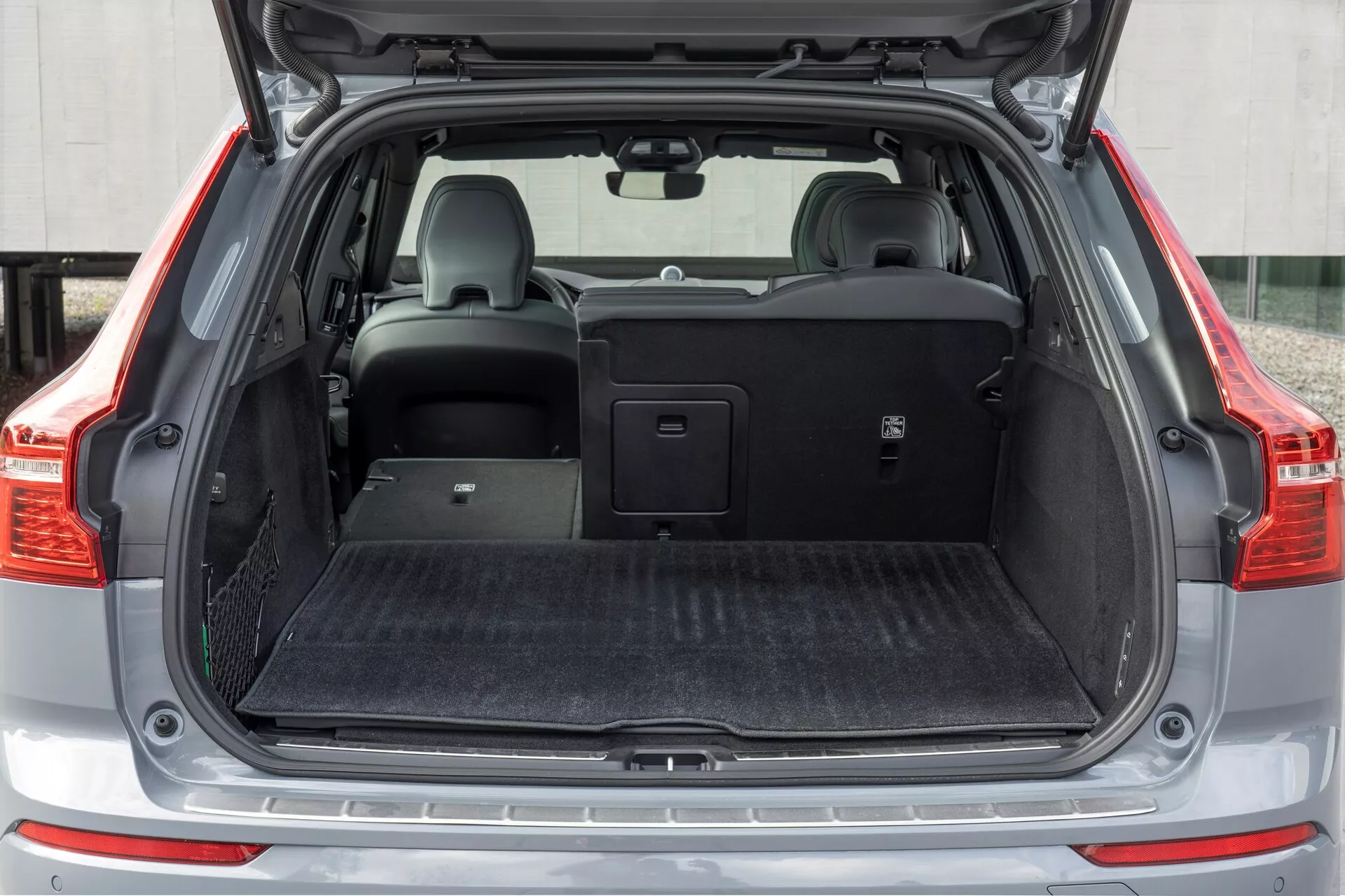Obraz przedstawia tylną przestrzeń ładunkową Volvo XC60 2021 (facelifting drugiej generacji), widzianą z prostopadłego kąta z otwartym bagażnikiem. Ujęcie średnie oddaje całą szerokość przestrzeni ładunkowej, z płaską podłogą pokrytą czarnym materiałem przypominającym dywan. Po lewej stronie znajduje się mały schowek z siatką. Tylne prawe siedzenie jest złożone, ukazując możliwość dzielenia składania, podczas gdy lewe siedzenie pozostaje w pozycji pionowej z zagłówkiem. Nad tylnymi siedzeniami umieszczona jest zasłona bagażnika, przedstawiająca wszechstronność przechowywania pojazdu.