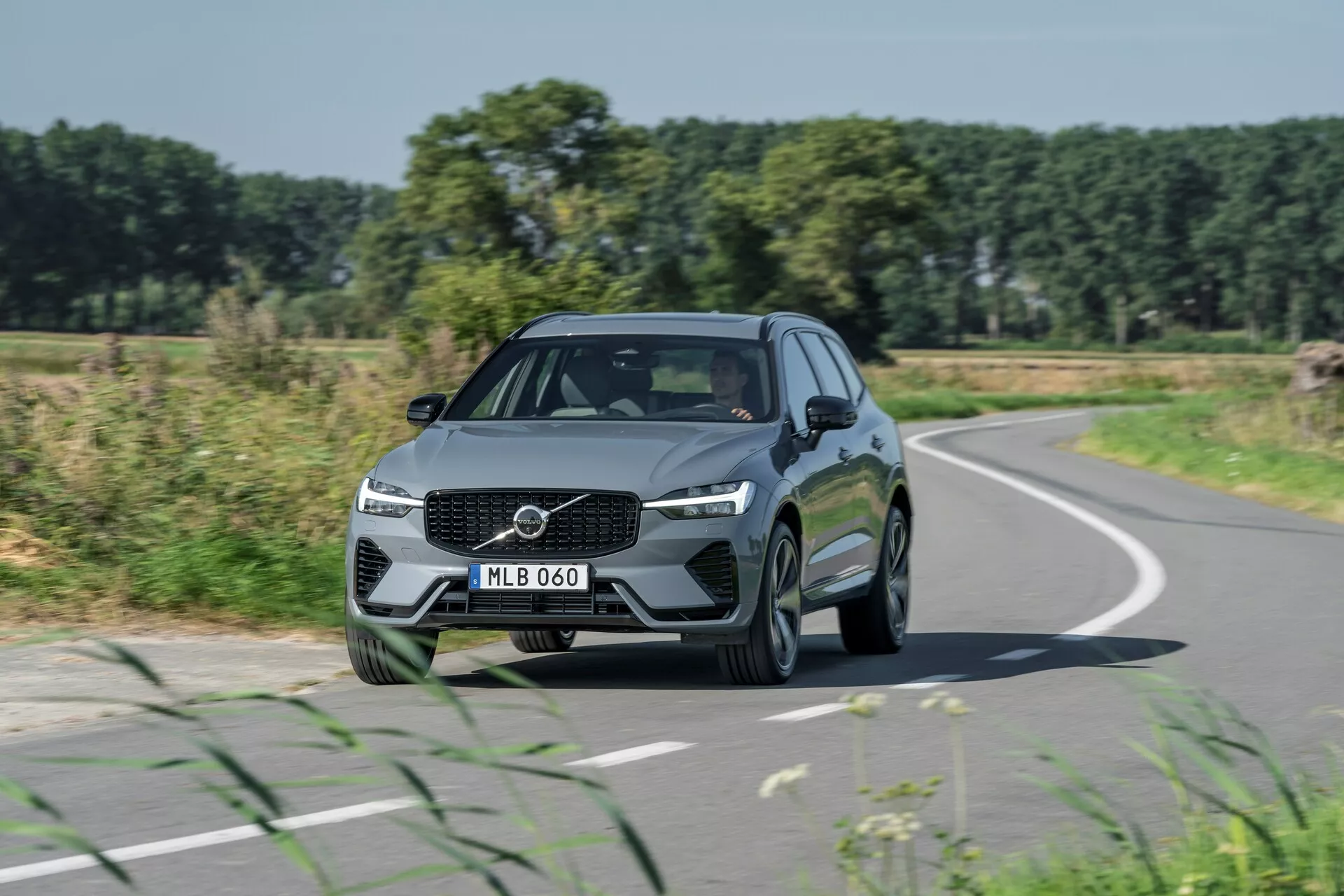 Obraz przedstawia Volvo XC60 2021 (odświeżenie 2. generacji) w ruchu z przedniego kąta trzy czwarte, podkreślając elementy designu takie jak wyrazista osłona chłodnicy z centralnie umieszczonym logo, kątowe reflektory LED i eleganckie linie maski. Samochód, poruszający się po lekko zakrzywionej drodze, ma metaliczne wykończenie i wieloramienne felgi aluminiowe, otoczone konturowanymi liniami i zintegrowanymi lusterkami bocznymi z kierunkowskazami. Na tle pól, drzew i jasnego błękitnego nieba, to przedstawienie podkreśla projektowanie samochodów, podróże drogowe i rzeczywiste scenariusze jazdy.