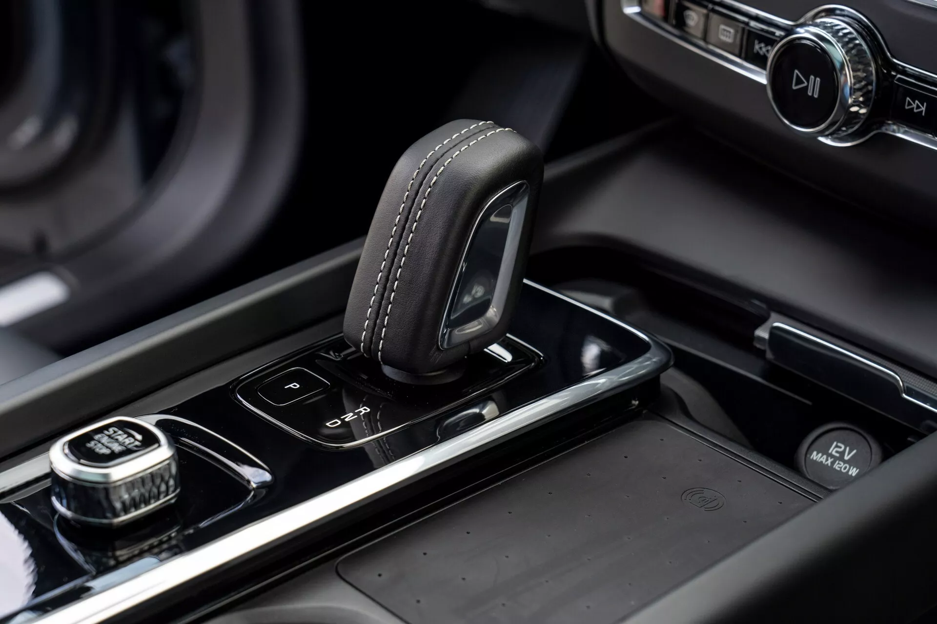 Zbliżenie konsoli środkowej w Volvo XC60 z 2021 roku (2. generacja, facelifting). Obraz podkreśla selektor biegów z wierzchem pokrytym skórą i detalem szwów, umieszczony na błyszczącej czarnej powierzchni. Po prawej stronie znajduje się pokrętło o ząbkowanej krawędzi, prawdopodobnie do nawigacji w systemie infotainment, z przyciskiem do wyboru przez naciśnięcie. Obok selektora biegów znajdują się przyciski do czujników parkowania i systemu start/stop. Konsola środkowa ma metalowe wykończenia, a w rozmytym tle znajduje się podświetlony ekran systemu infotainment, podkreślając nowoczesny, wysokiej jakości design pojazdu i jego rzemiosło.