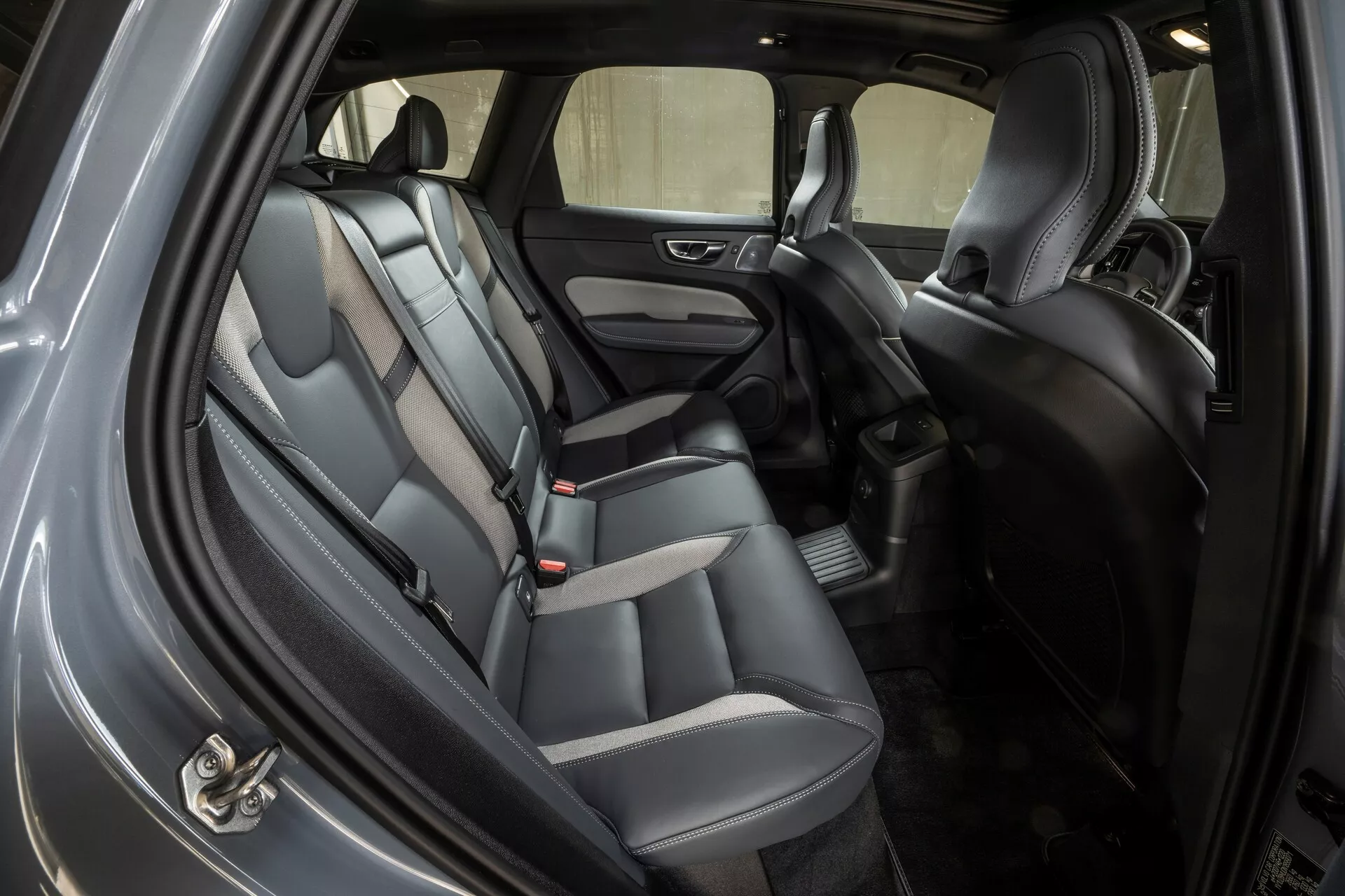 Ujęcie wnętrza Volvo XC60 z 2021 roku, ukazujące tylną przestrzeń siedzeń pod kątem ukośnym z otwartymi tylnymi drzwiami od strony krawężnika. Samochód wyposażony jest w dwukolorowe siedzenia skórzane lub ze skóry ekologicznej, zagłówki, sprzączki pasów bezpieczeństwa dla trzech pasażerów oraz tapicerowane panele drzwi z wbudowanym głośnikiem i sterowaniem elektrycznych szyb. Podłoga jest pokryta dywanikiem z dodatkową matą, a widoczna między przednimi siedzeniami konsola środkowa zawiera przestrzeń do przechowywania. Dobrze oświetlona kabina sugeruje oświetlenie ambientowe i sufitowe, oferując dobrą widoczność przez boczne i tylne okna.