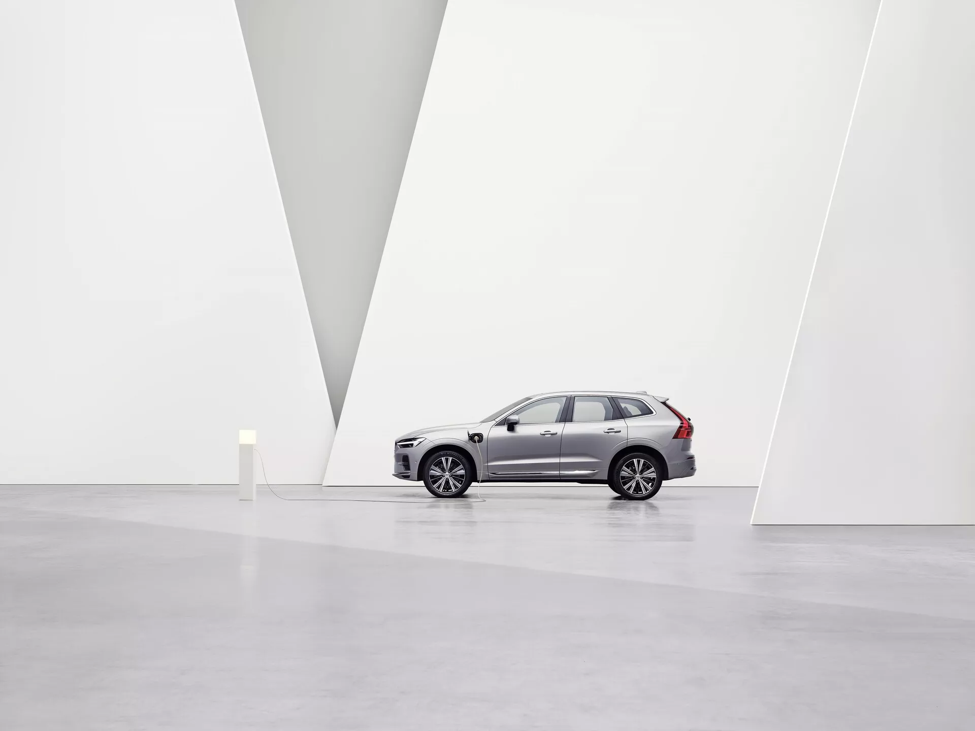 Obraz przedstawia samochód SUV Volvo XC60 z 2021 roku drugiej generacji po faceliftingu w przestronnej, minimalistycznej galerii sztuki z białymi ścianami i lśniącą podłogą. Pojazd widoczny z profilu na lewo podkreśla aluminiowe felgi, wyraźne linie boczne, lusterka boczne, przyciemniane szyby oraz charakterystyczne relingi dachowe. Z lekkiego niskiego kąta widać przedni grill, reflektory LED, zintegrowane kierunkowskazy oraz czujniki parkowania na przednim zderzaku. Czyste otoczenie i przegroda w kształcie litery V w ścianie podkreślają design samochodu i jego pozycję premium na rynku w miękkim, rozproszonym oświetleniu.