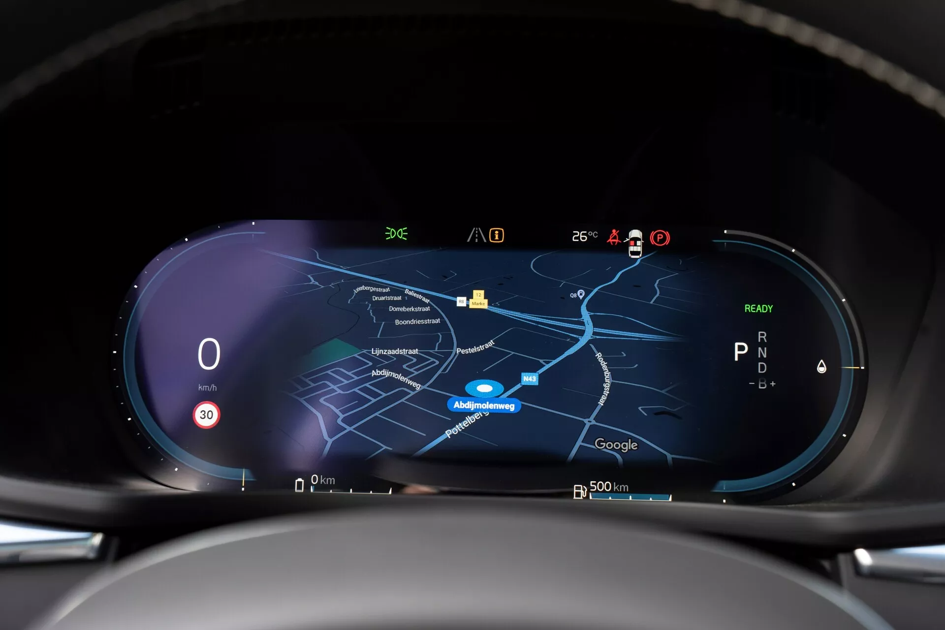 Widok wnętrza Volvo XC60 2021 drugiej generacji po liftingu, ukazujący wysokiej rozdzielczości cyfrowy zestaw wskaźników z perspektywy kierowcy. Zestaw wskaźników zawiera szerokoekranowy wyświetlacz z prędkościomierzem wskazującym zero, wskaźnikiem biegu pokazującym 'P' oraz centralną mapą nawigacyjną w trybie nocnym. Różne ikony dla odbioru sygnału, temperatury zewnętrznej, naładowania baterii i funkcji wspomagania kierowcy są widoczne na górnej krawędzi, wskazując na łączność w czasie rzeczywistym i zaawansowane możliwości technologiczne.