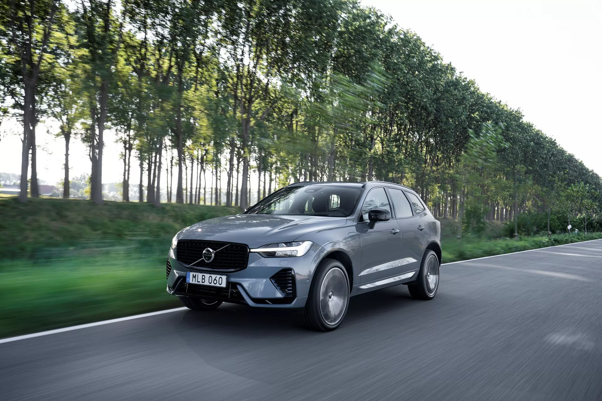 Na obrazie przedstawiono poruszające się Volvo XC60 2021 (lifting 2. generacji) uchwycone pod ujęciem trzech czwartych z przodu, ukazujące stronę kierowcy samochodu. Ten nowoczesny luksusowy SUV charakteryzuje się eleganckim designem i widocznym emblematem na przedniej kracie. Zdjęcie zrobione pod niewielkim kątem w dół pokazuje maskę, zderzak, kratę, reflektory i koła. Ciemny metaliczny pojazd, prawdopodobnie szary lub czarny, z błyszczącym wykończeniem, posiada koła ze stopu, reflektory LED i aerodynamiczne nadwozie. Rozmyte tło ulicy z drzewami sugeruje ruch, podkreślając możliwości dynamiczne samochodu.