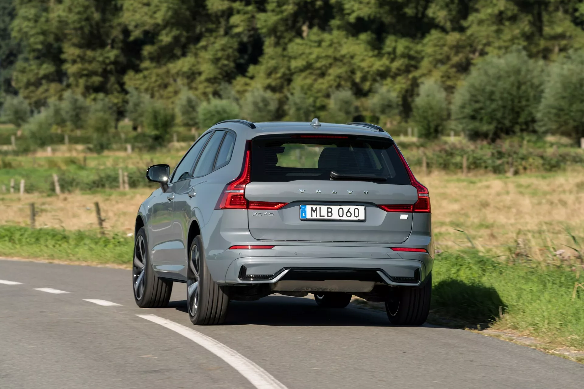 Tylny widok trzech-czwartych Volvo XC60 2021 poruszającego się po asfaltowej drodze. Uchwycone z podwyższonego kąta, to nowoczesne SUV prezentuje swoją błyszczącą szarą farbę, charakterystyczne tylne światła LED, podwójne końcówki wydechu i tylny dyfuzor. Na rzeźbionej klapie bagażnika widoczne są emblemat producenta i oznaczenie modelu, uzupełnione przyciemnianymi szybami i aluminiowymi felgami o wieloramiennym wzorze, sugerującymi sportową wersję. Obraz dostarcza wyraźnego wglądu w aerodynamiczny design i wysokiej klasy funkcje charakterystyczne dla nowoczesnego pojazdu.