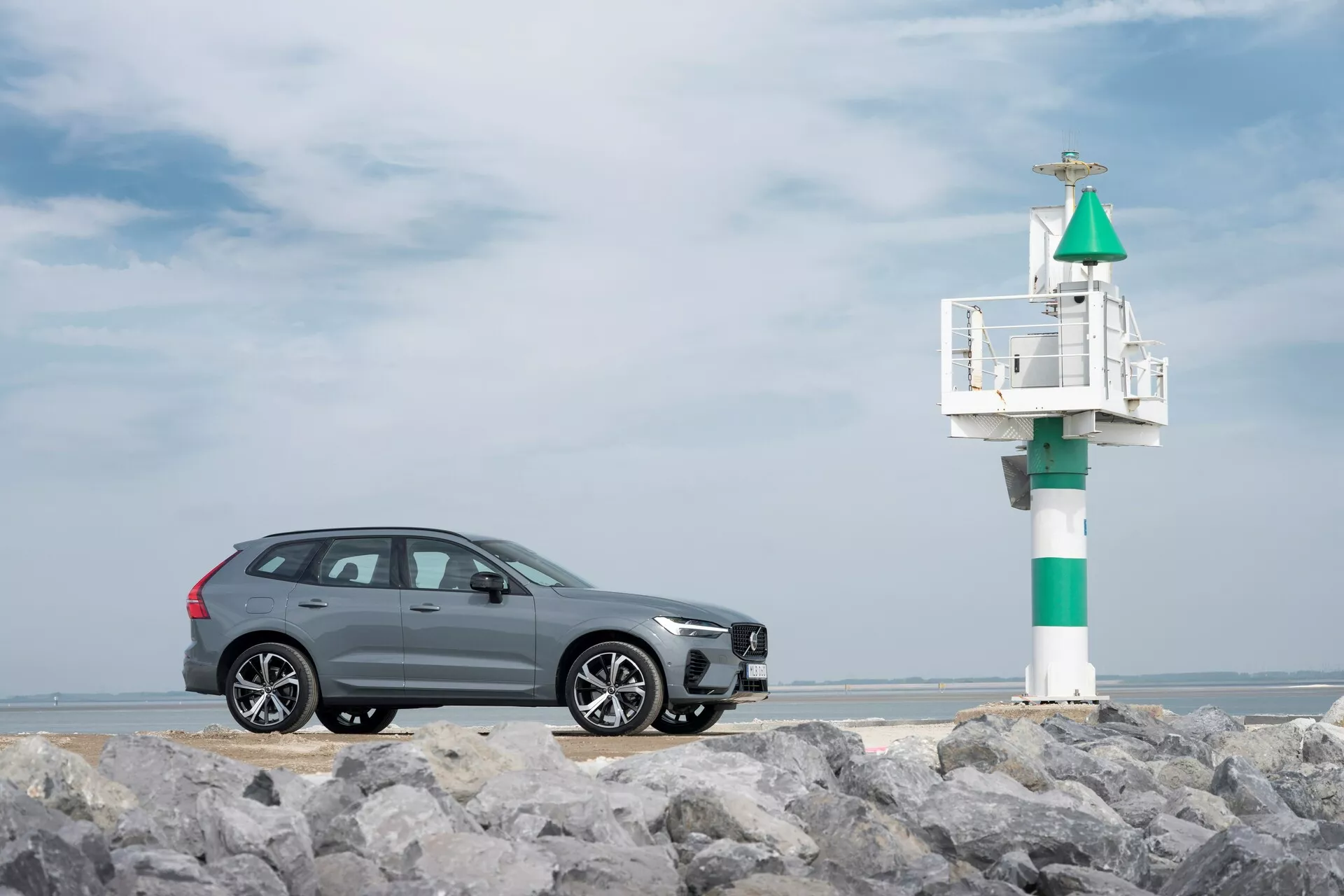 Widok z profilu szarego SUV-a Volvo XC60 2021 (druga generacja, lifting) na skalistym podłożu z morskim sygnalizatorem w tle. Elegancki design obejmuje chromowane listwy okienne, wieloramienne felgi aluminiowe i zintegrowany kierunkowskaz w lusterku bocznym, uchwycony na tle częściowo zachmurzonego wybrzeżowego nieba.