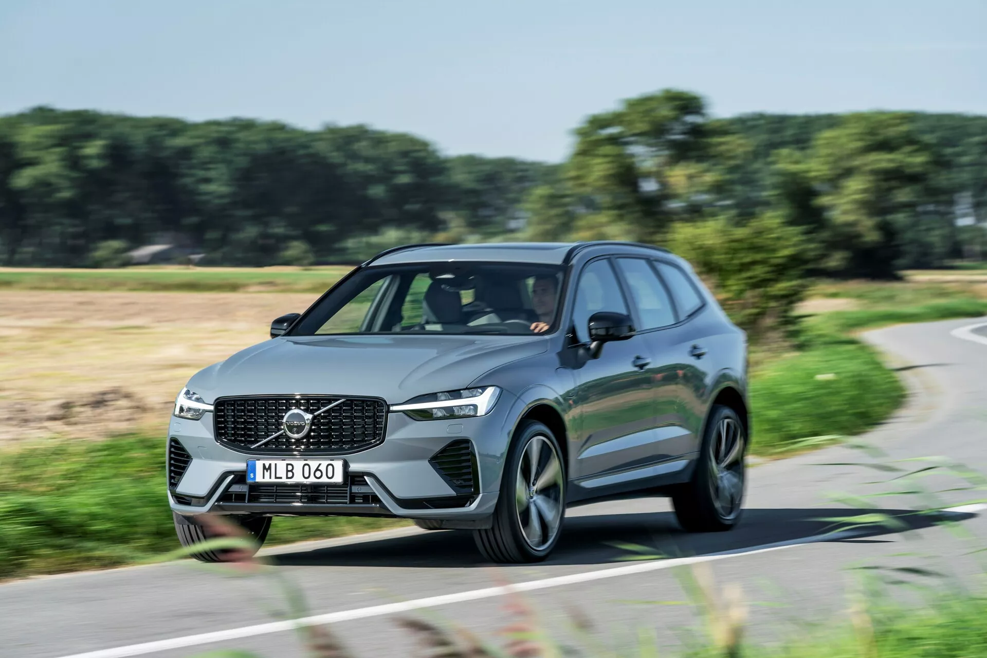 Dynamiczne zdjęcie w świetle dziennym ruchomego Volvo XC60 2021 (druga generacja-facelift) SUV uchwyconego z kąta trzech czwartych przedniego. Obrazek pokazuje przedni grill z wyraźnym emblematem Volvo, kątowe reflektory LED, linie aerodynamiczne i nowoczesne wieloramienne felgi aluminiowe. Światła do jazdy dziennej są włączone, co wskazuje, że samochód jest w ruchu. Zdjęcie podkreśla elegancki design i obecność na drodze tego nowoczesnego rodzinnego lub luksusowego pojazdu użytkowego.