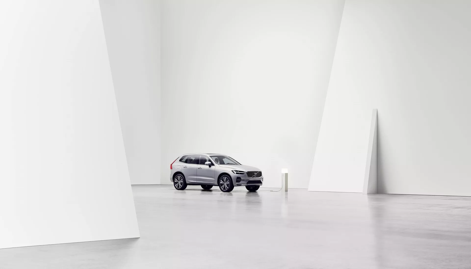 Obraz Volvo XC60 2021 (2. generacja, facelift) umieszczonego centralnie w minimalistycznym, przestronnym, dobrze oświetlonym środowisku studyjnym, uchwyconego z przedniej perspektywy trzech czwartych. Jasno srebrny odcień samochodu kontrastuje z ciemniejszymi kołami i częścią przedniego grilla, pokazując nowoczesne elementy designu SUV-a, takie jak zakrzywione i kanciaste linie. Widok podkreśla przedni grill, reflektory, maskę, przednią szybę oraz stronę kierowcy, w tym przednie i tylne drzwi oraz boczne okna. Starkowe, białe tło akcentuje samochód, koncentrując uwagę na jego designie i cechach, odpowiedni do celów promocyjnych lub artystycznych.