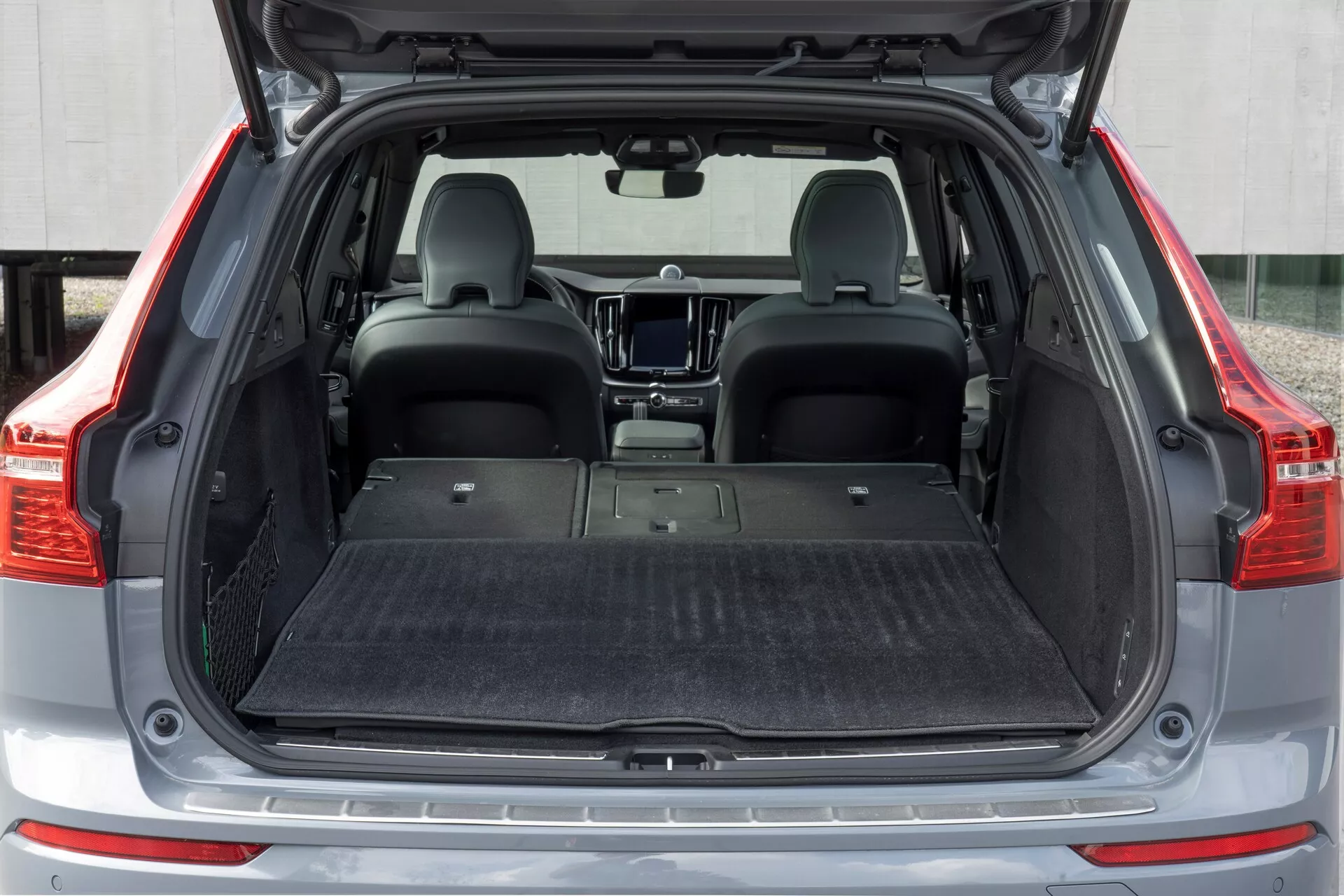 Tylny obszar ładunkowy Volvo XC60 2021 (druga generacja facelift) widziany z prostego tylnego perspektywy. Otwarta przestrzeń bagażnika ujawnia pustą, wyściełaną dywanem płaską podłogę bagażnika, z złożonymi tylnymi siedzeniami w celu zwiększenia pojemności ładunkowej. Wnętrze jest nowoczesne, z matą ochronną na podłodze, ale bez widocznych narzędzi organizacyjnych, takich jak siatki bagażowe lub haki.