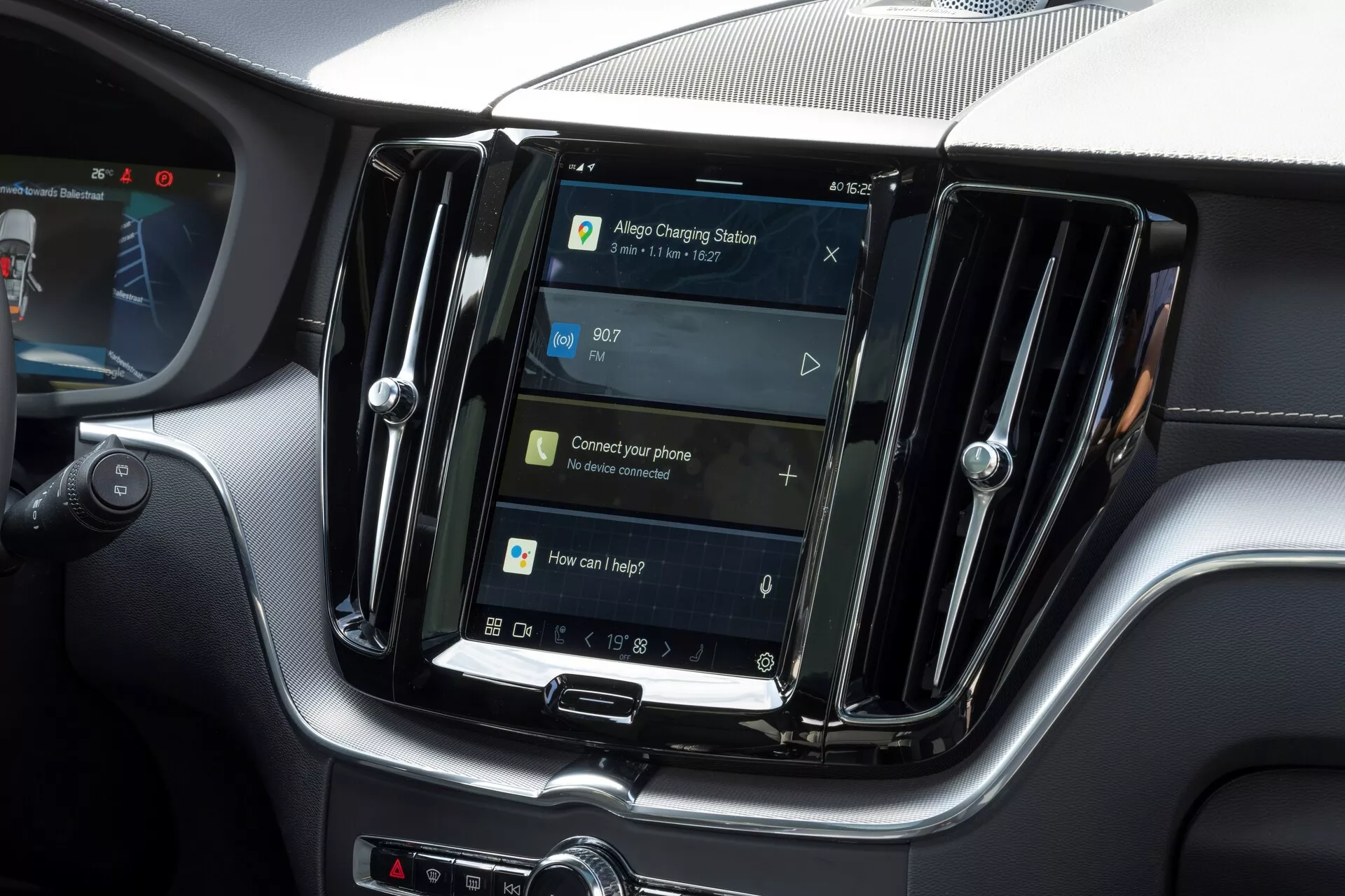 Widok wnętrza Volvo XC60 2021 (druga generacja, lifting) z naciskiem na centralny system infotainment. Wyświetlacz ustawiony pionowo, podobny do tabletów i smartfonów, jest otoczony metalowymi wentylatorami, co sugeruje wyższy model. Poniżej ekranu znajdują się fizyczne przyciski i pokrętło obrotowe, co sugeruje połączenie dotykowego i fizycznego sterowania. Ekran nawigacyjny wskazuje na funkcję GPS, z monitami dotyczącymi podłączenia telefonu i sterowania głosem, sugerując funkcje takie jak Bluetooth, rozpoznawanie głosu i integrację smartfona. Czas jest wyświetlany o godzinie 09:07. Obraz wyróżnia cechy systemu infotainment bez pokazywania logo lub identyfikatorów modelu.
