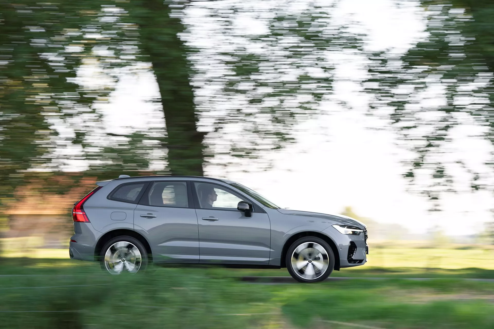 Obraz przedstawia SUV-a Volvo XC60 2021 (druga generacja, facelift) w ruchu, uchwyconego techniką panningu, która rozmywa tło, zachowując ostrość samochodu. Widoczny z boku kierowcy w pełnym profilu bocznym, metalicznie szary pojazd ma pięć drzwi, aluminiowe felgi i standardowe opony drogowe. Scena sugeruje jazdę dzienną przez naturalne otoczenie, z rozmytymi drzewami i trawą w tle, podkreślając ruch samochodu.