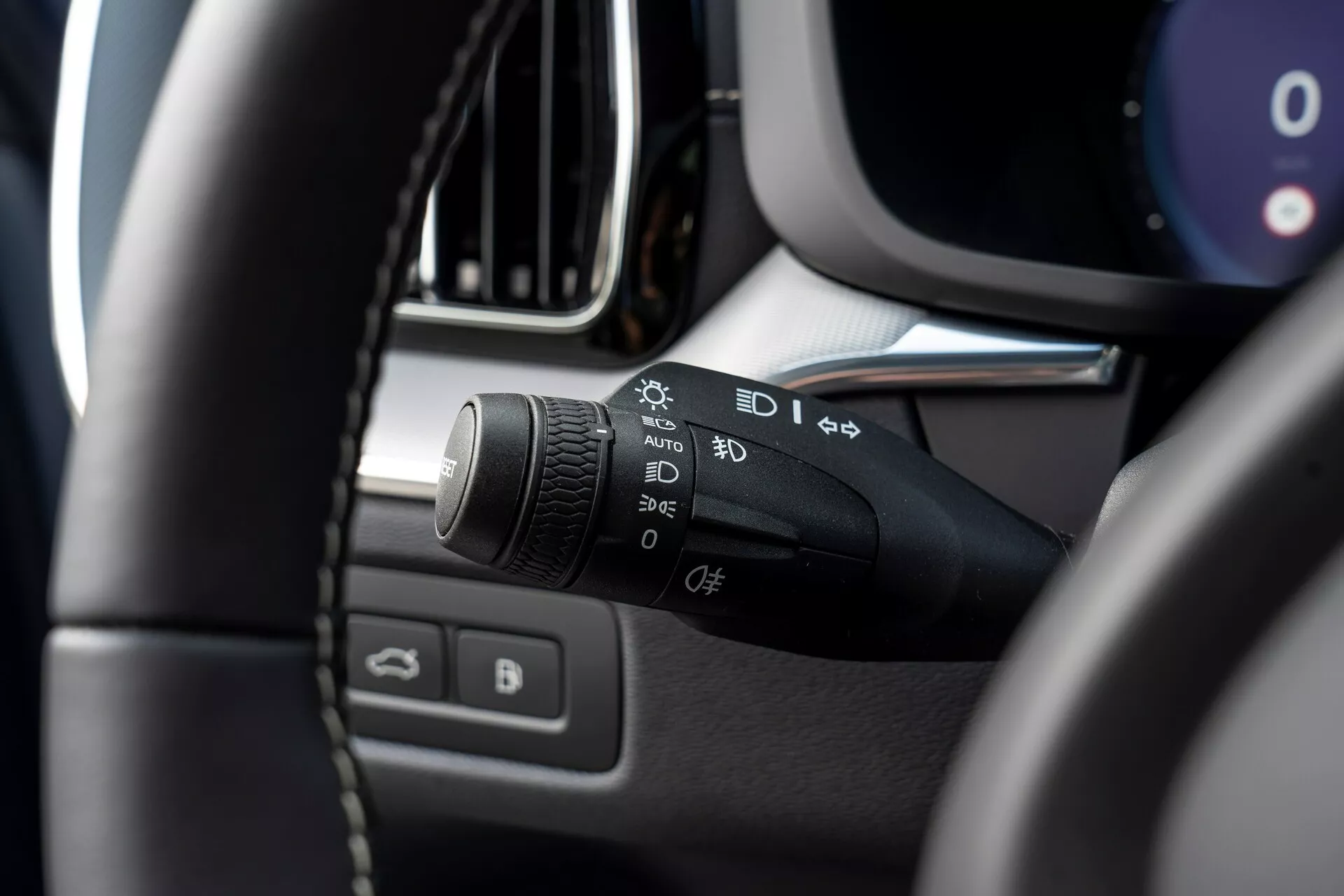 Zbliżenie wnętrza Volvo XC60 2021 (druga generacja, facelifting) z focusowaniem na lewej kolumnie kierowniczej. Głównym tematem jest dźwignia sterująca reflektorami i kierunkowskazami, z symbolami dla świateł automatycznych, świateł postojowych, reflektorów, ustawień świateł przeciwmgielnych oraz funkcji personalizowanej. Obraz zrobiony z siedzenia kierowcy, lekko w górę i na lewo, pokazujący rozmyty lewy górny róg kierownicy na pierwszym planie, podkreślając dźwignię sterowania. Dodatkowe rozmyte szczegóły obejmują części panelu instrumentów, konsoli centralnej, krawędź kratki wentylacyjnej lub ekranu infotainmentu oraz drzwi kierowcy z przyciskami do sterowania oknami i lusterkami.