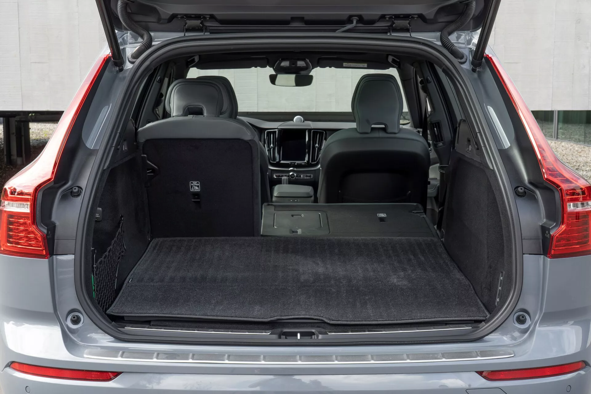 Zbliżenie tył widok na 2021 Volvo XC60 drugiej generacji po liftingu, przedstawiający otwarty bagażnik pod niskim kątem. Obraz ukazuje przestronną przestrzeń bagażową z minimalnym naciskiem na nadkola, wysuwaną osłonę bagażnika i wykończenie wnętrza wykładziną mające na celu redukcję hałasu i ochronę. Częściowo złożone tylne siedzenia demonstrują wszechstronność, podczas gdy widoczna płyta ochronna na progu załadunku, czujniki parkowania na zderzaku tylnym, oświetlenie LED wewnątrz bagażnika i mocowania do przewozu towarów podkreślają funkcjonalność i dbałość o szczegóły.